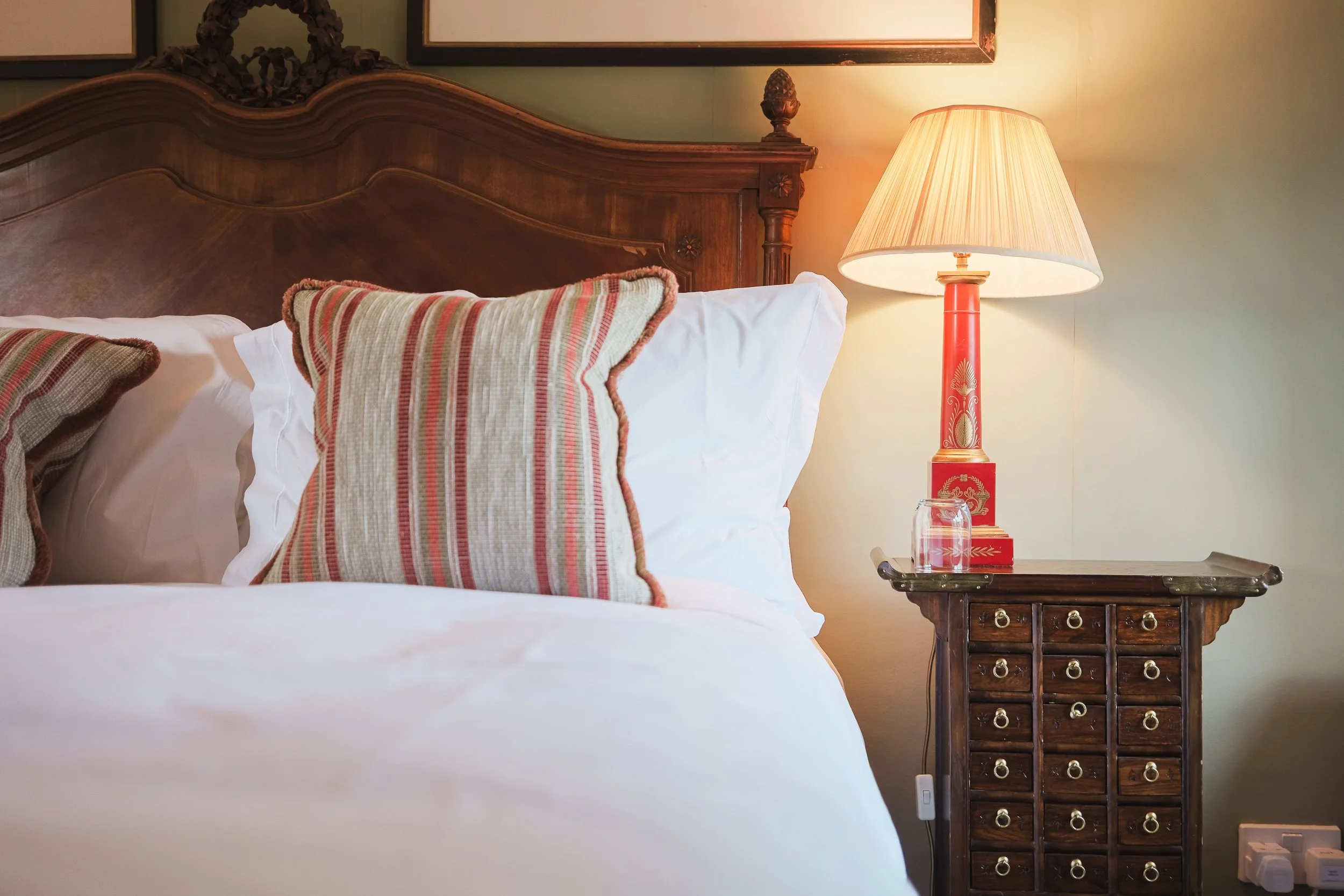 A bedroom with a wooden headboard, decorative pillows with striped patterns, a bedside table with a red and gold lamp, and framed artwork on the wall.
