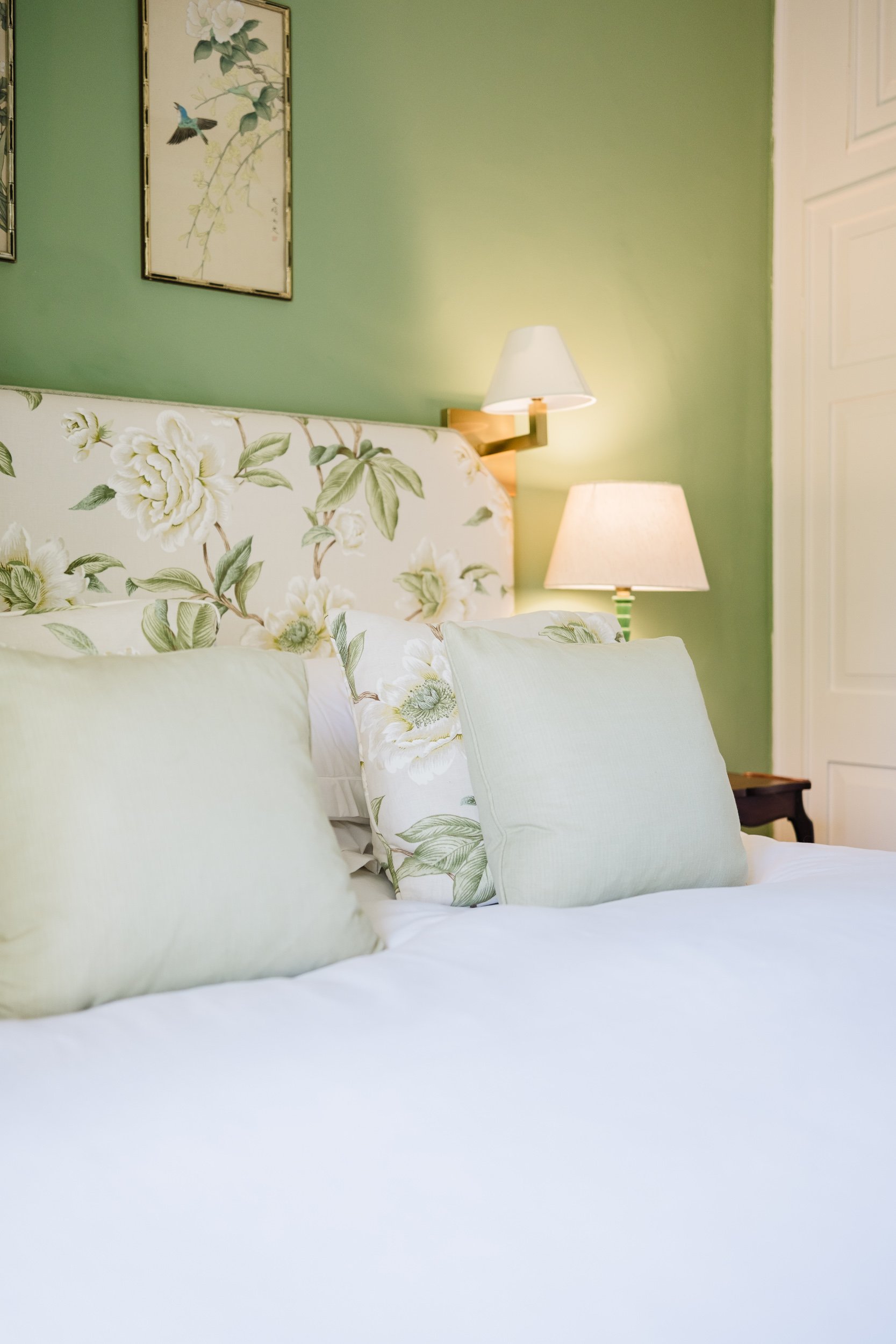 A neatly made bed with white floral patterned pillows and a headboard against a green wall, featuring two wall lamps. There is a framed picture of birds on the wall.