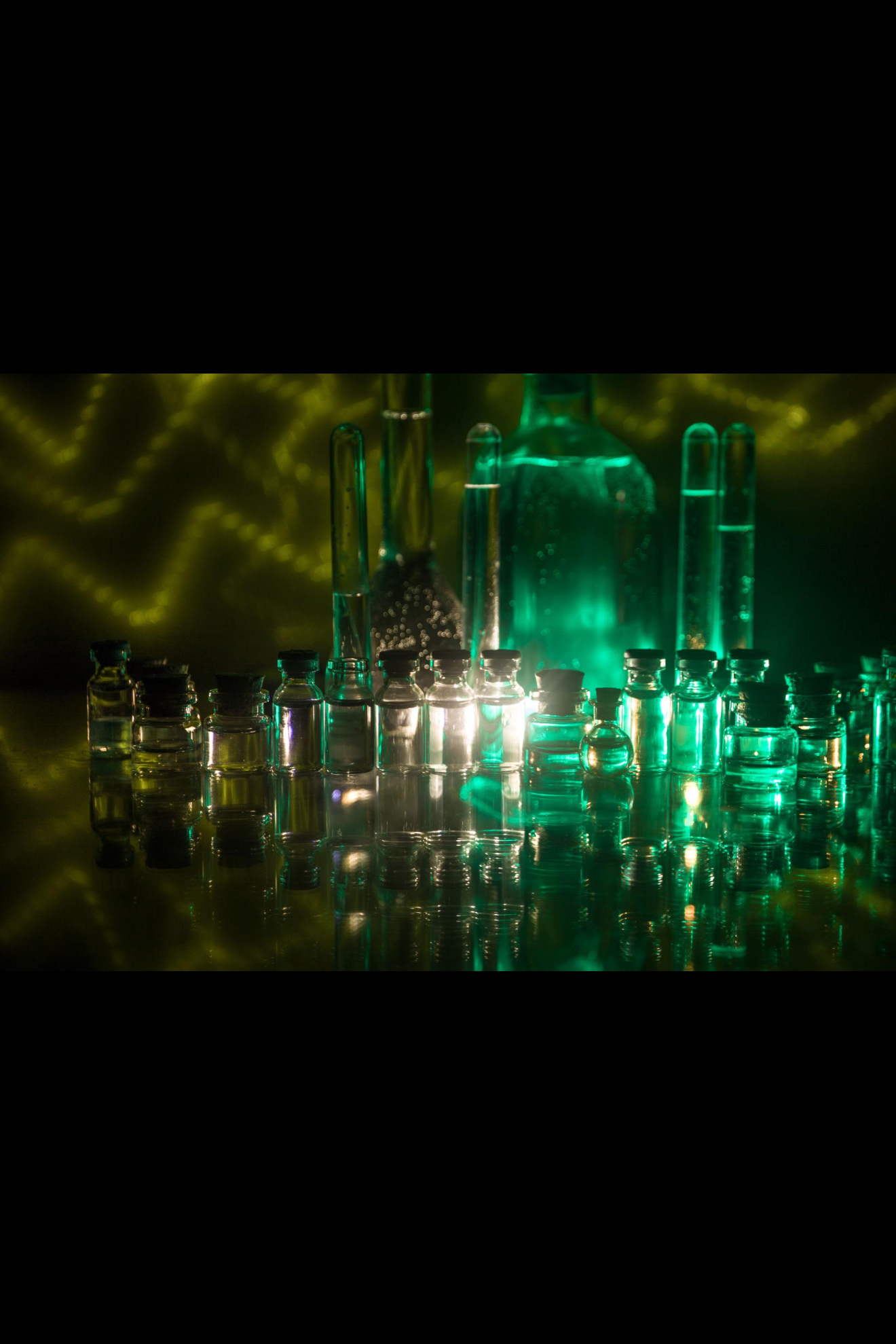 A collection of small glass vials with black caps, illuminated by green and yellow lighting, with larger glass containers and test tubes in the background, creating a laboratory or scientific setting.