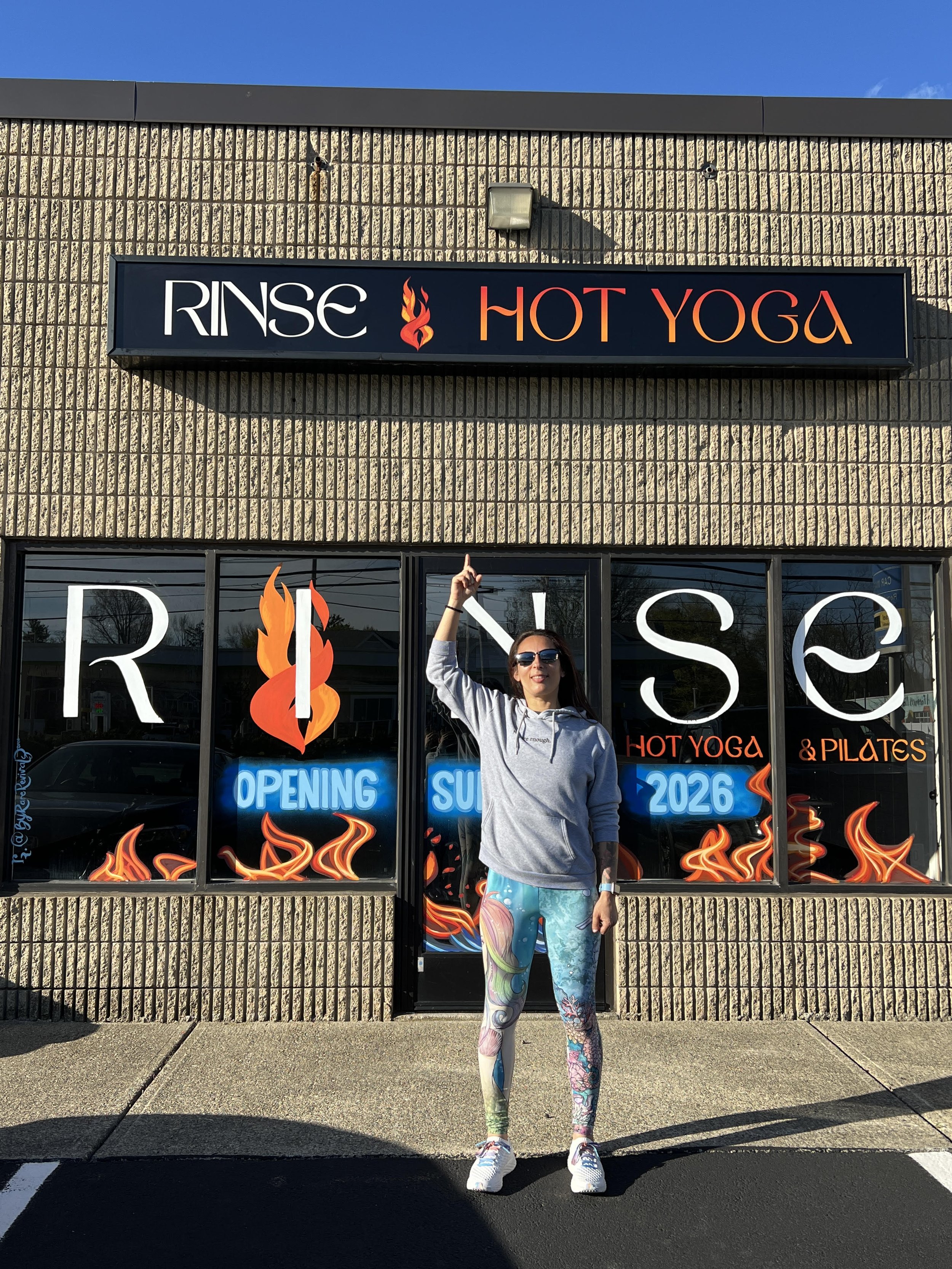 Stacy the studio owner is standing in front of the yoga studio pointing to the RINSE HOT YOGA sign on the building.