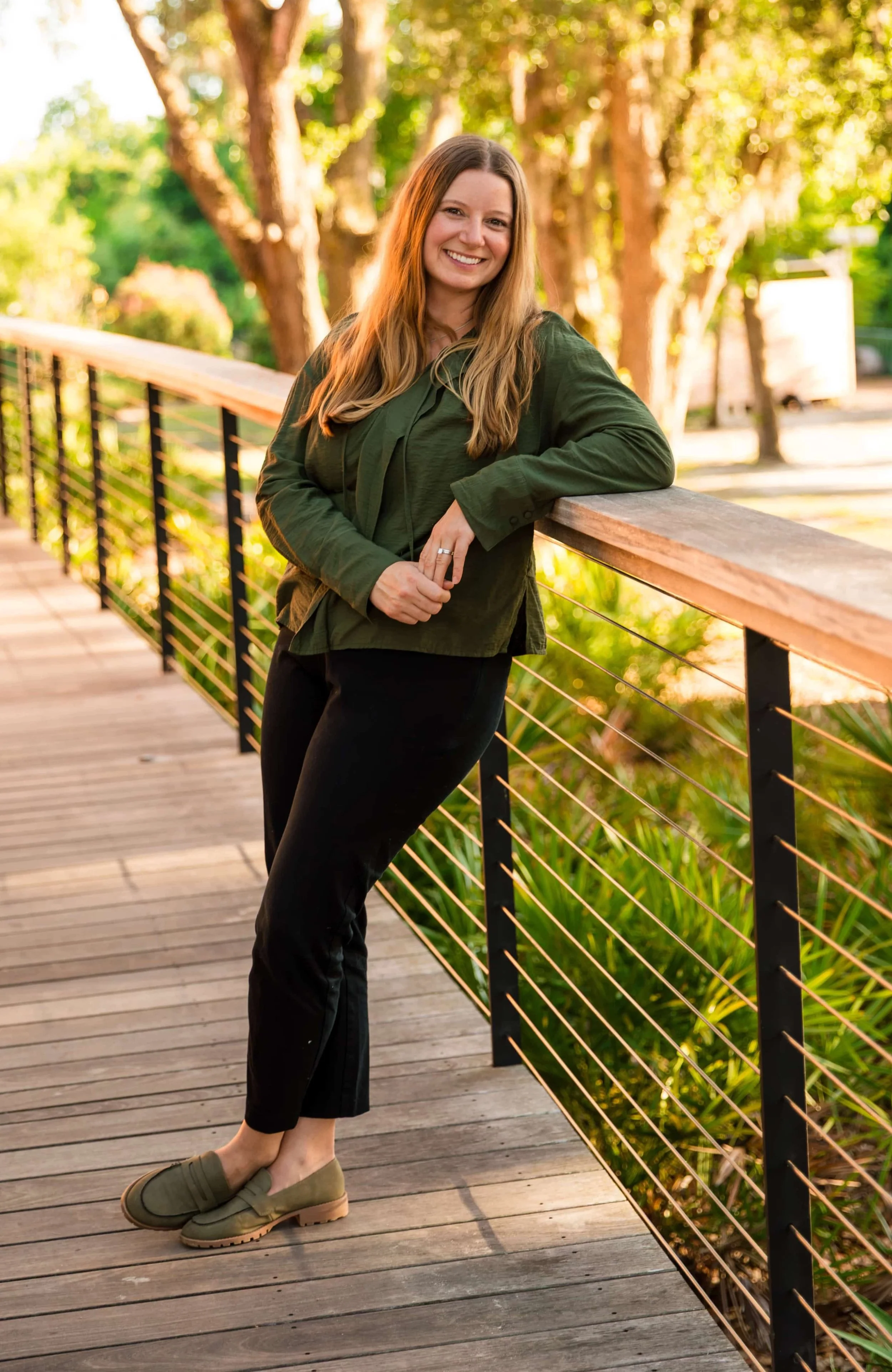 A woman with long blonde hair smiling and leaning on a wooden and metal railing on a walking bridge in a park during sunset, dressed in green jacket, black pants, and tan shoes.
