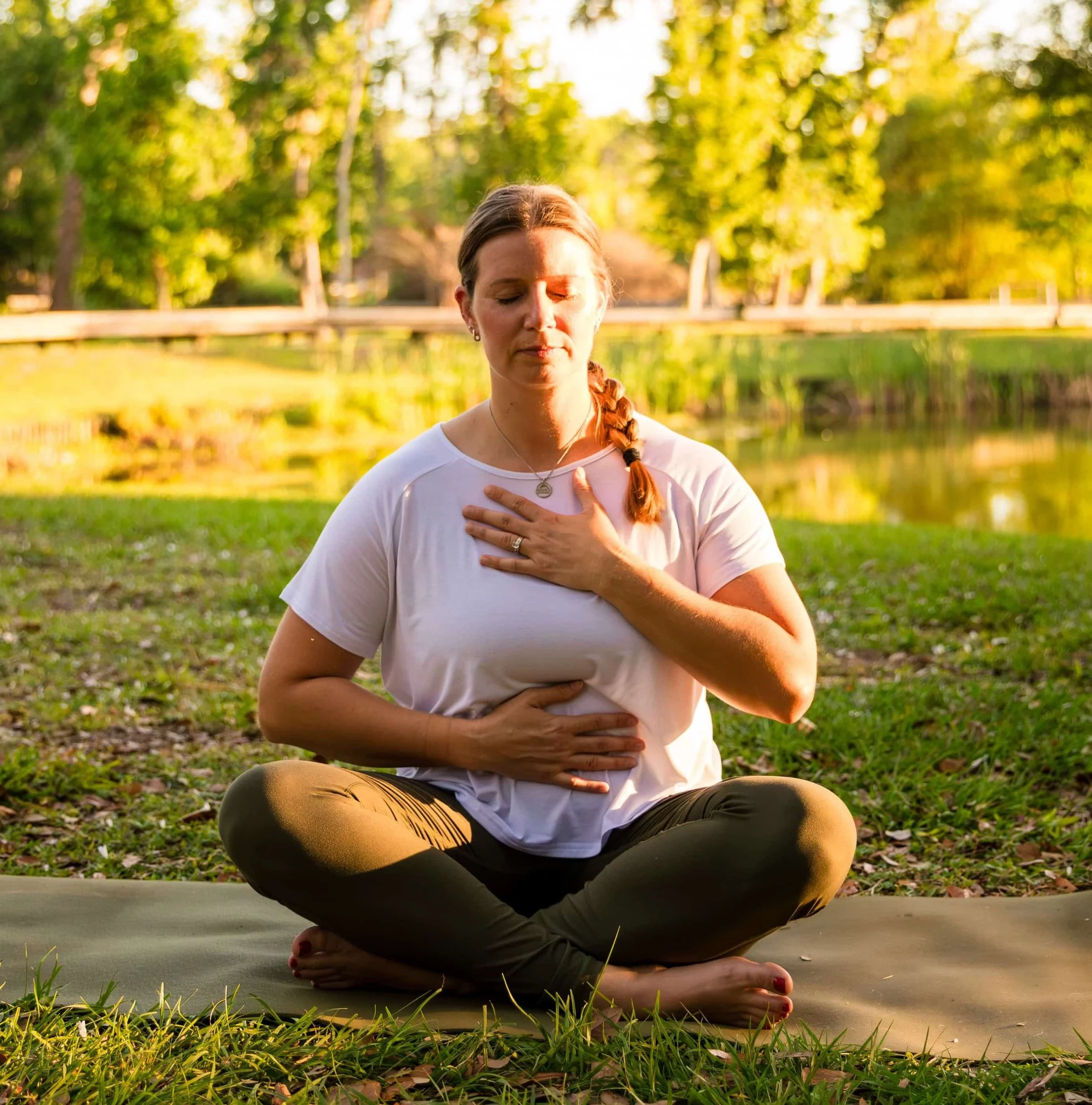 A woman practicing yoga outdoors on a gray mat, sitting in a meditative pose with her hands on her chest and stomach, eyes closed, surrounded by green trees and grass during sunset.