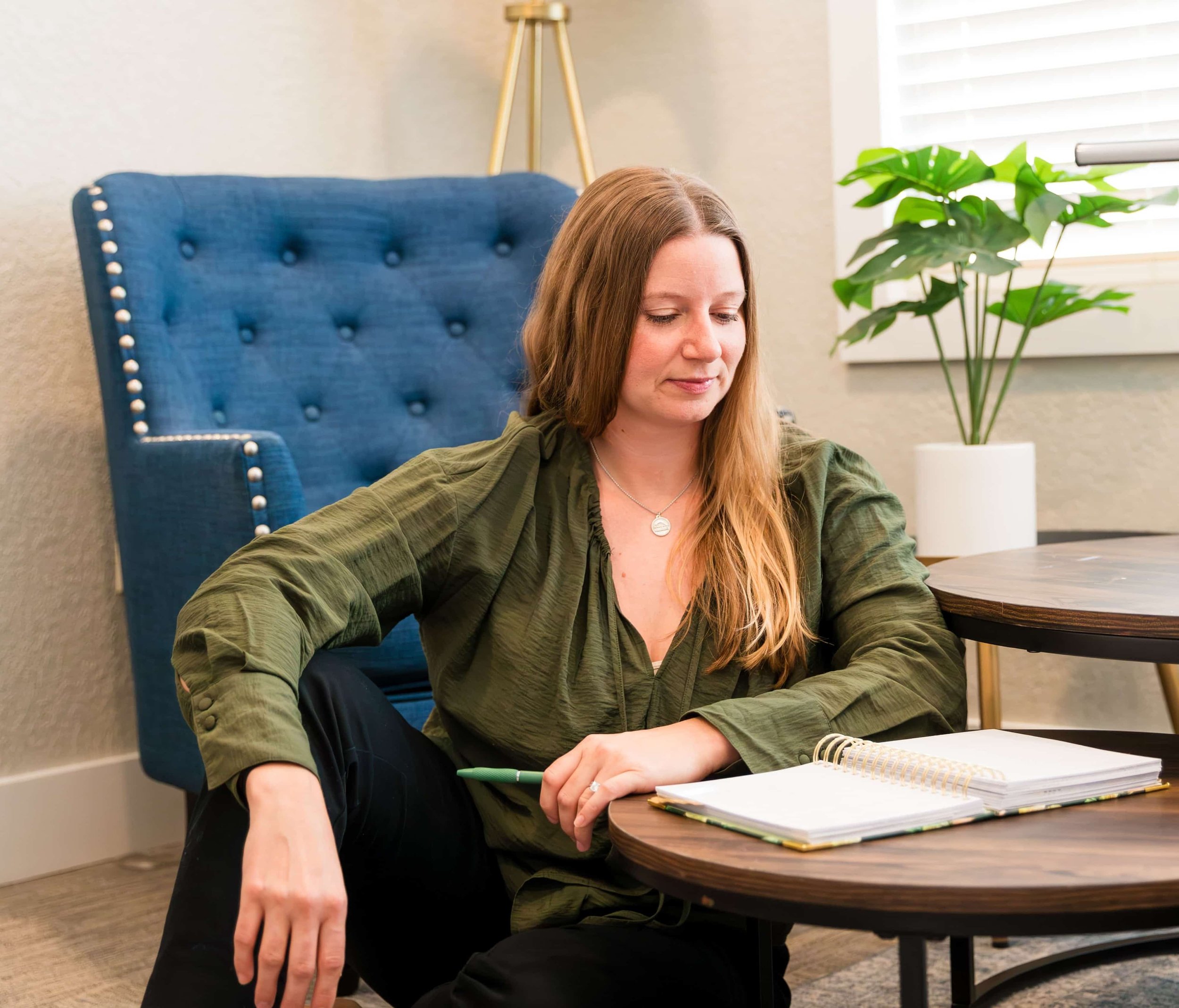 A woman with long brown hair sitting at a round wooden table in a room with beige walls, a large green plant in a white pot, and a blue upholstered chair with button tufting and nailhead trim. She is wearing a green jacket, a necklace, and has notebooks open in front of her, holding a green pen.