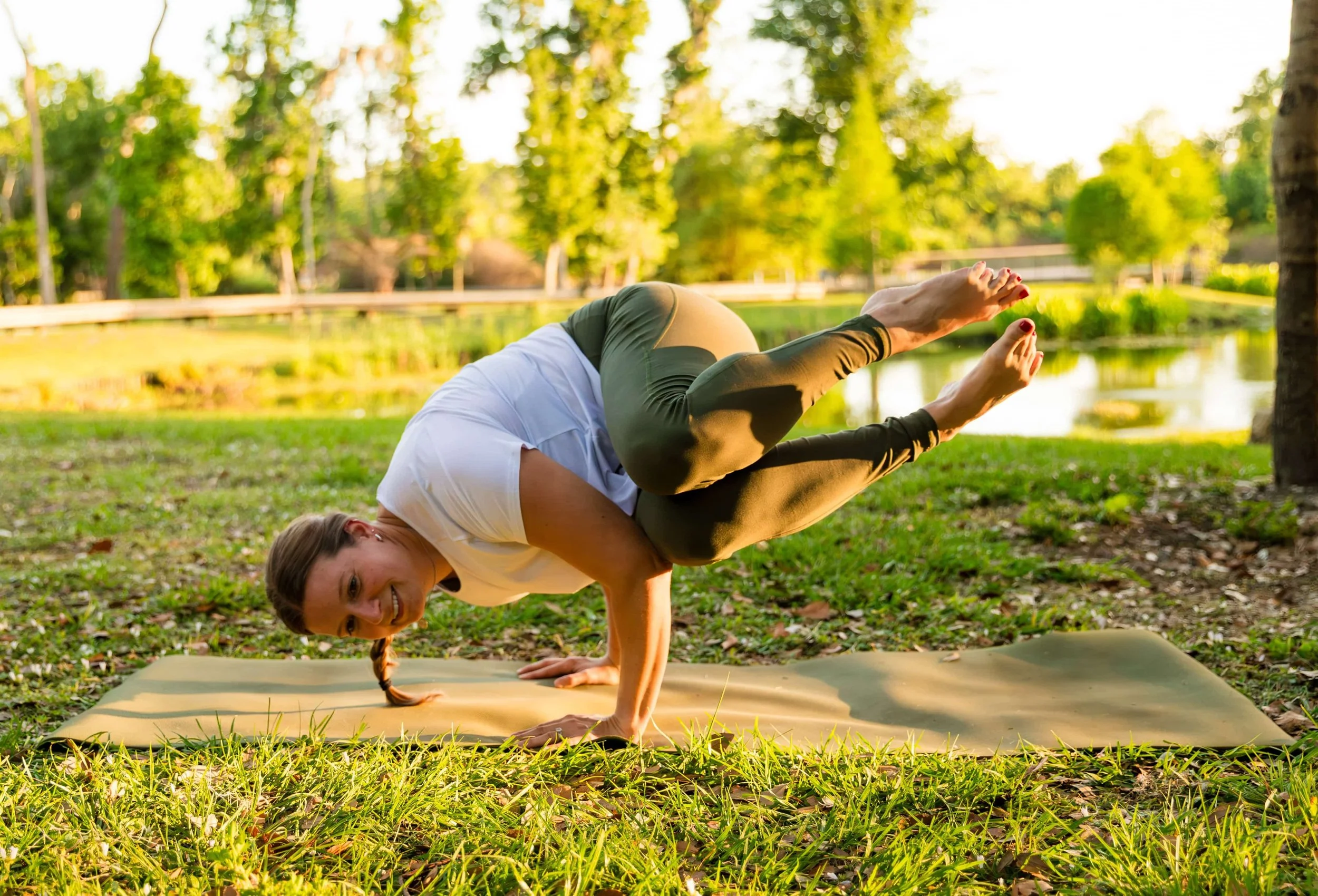 A woman doing yoga in a park during sunset, balancing on her hands with her legs bent over her back and her head turned to the side, surrounded by green trees and grass.