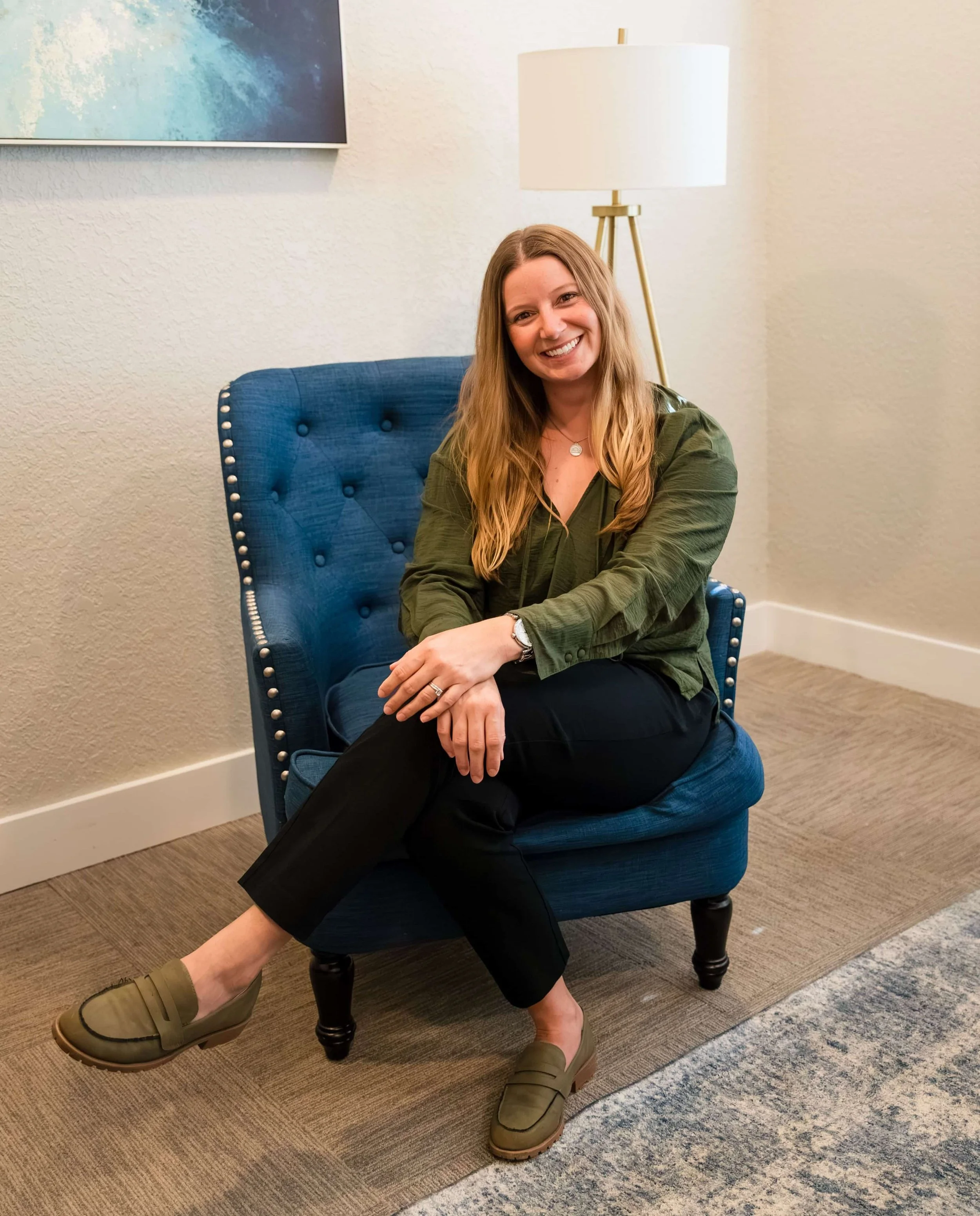 A woman with long blonde hair, smiling, sitting on a blue tufted armchair in a room with beige textured walls. She wears an olive green jacket, black pants, and tan slip-on shoes. A floor lamp and a painting are visible in the background.