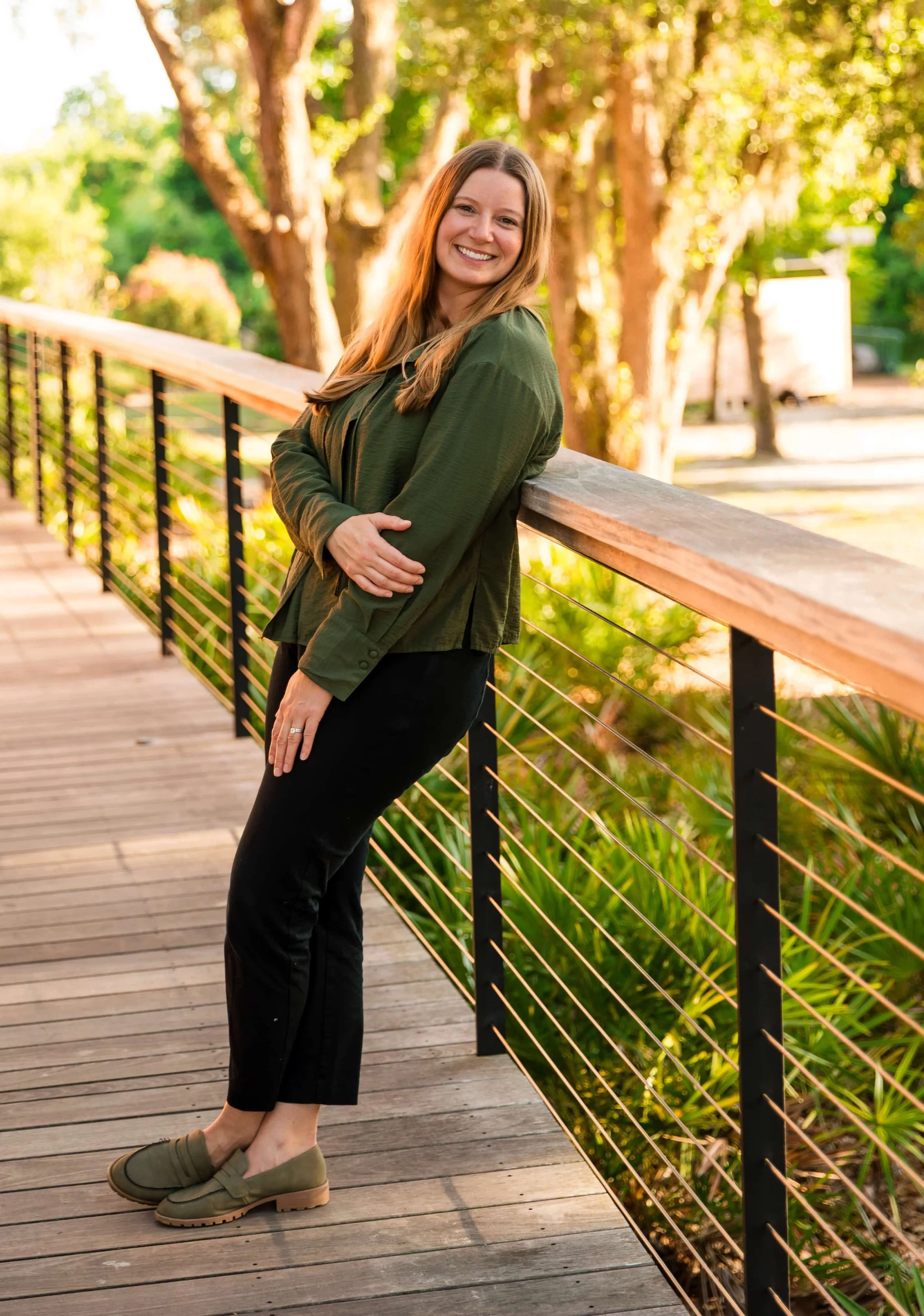 A woman with long blonde hair, wearing a dark green jacket, black pants, and green shoes, leaning against a railing on a wooden bridge, smiling outdoors with green trees and sunlight in the background.