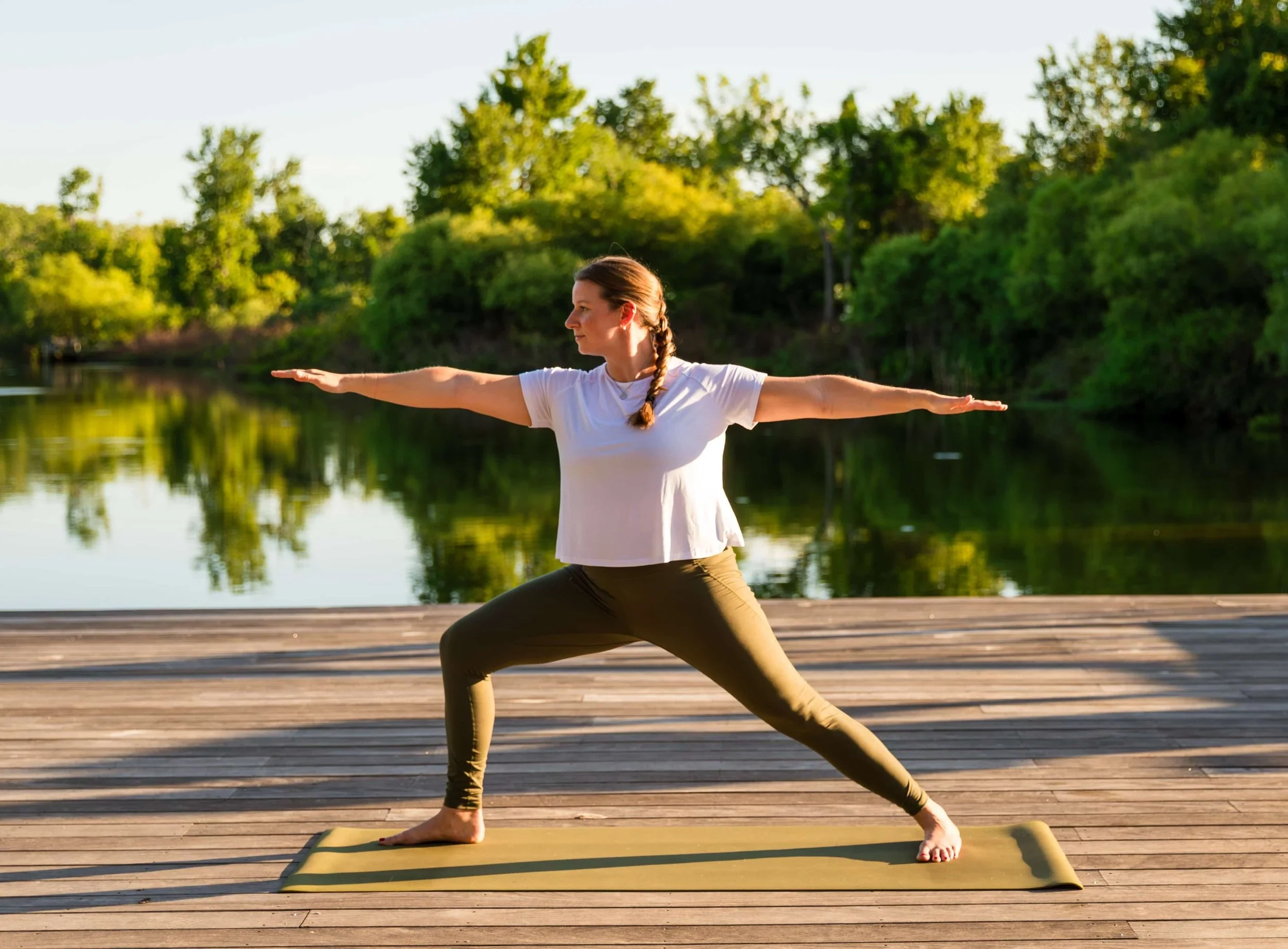A woman practicing yoga outdoors by a lake during sunset. She is in Warrior II pose on a yoga mat on a wooden deck, with trees and water in the background.