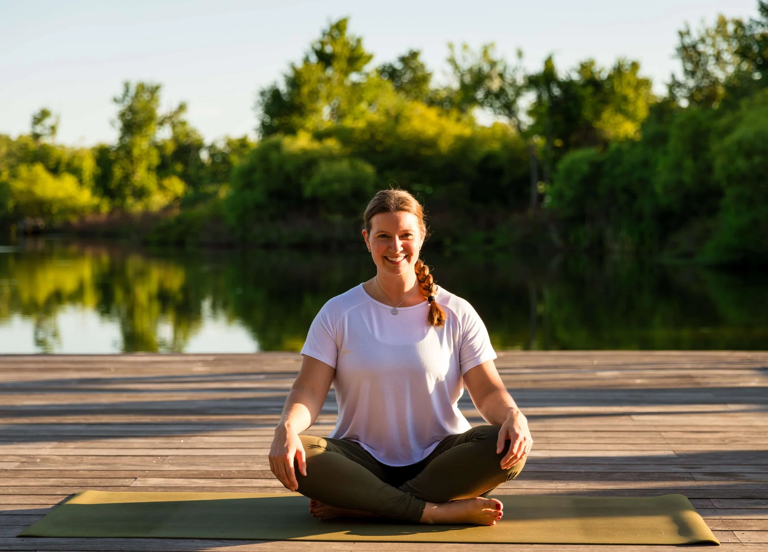 A woman practicing yoga outdoors on a wooden dock by a river, sitting in a cross-legged meditative pose with a smile, wearing a white t-shirt and dark pants, surrounded by greenery and trees in the background.