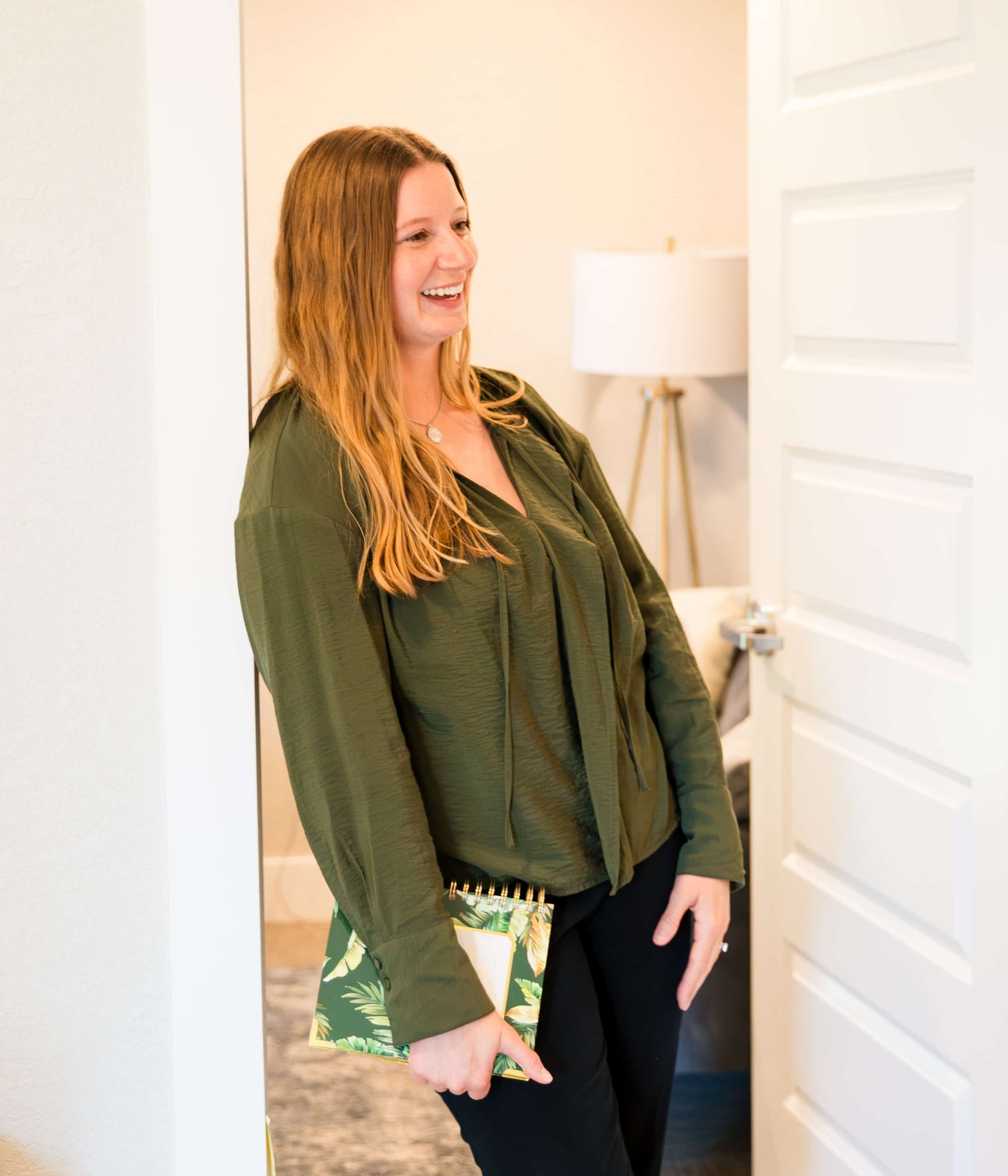 A woman with long red hair, smiling, holding a gift bag with a green and yellow tropical leaf pattern, standing in a doorway.