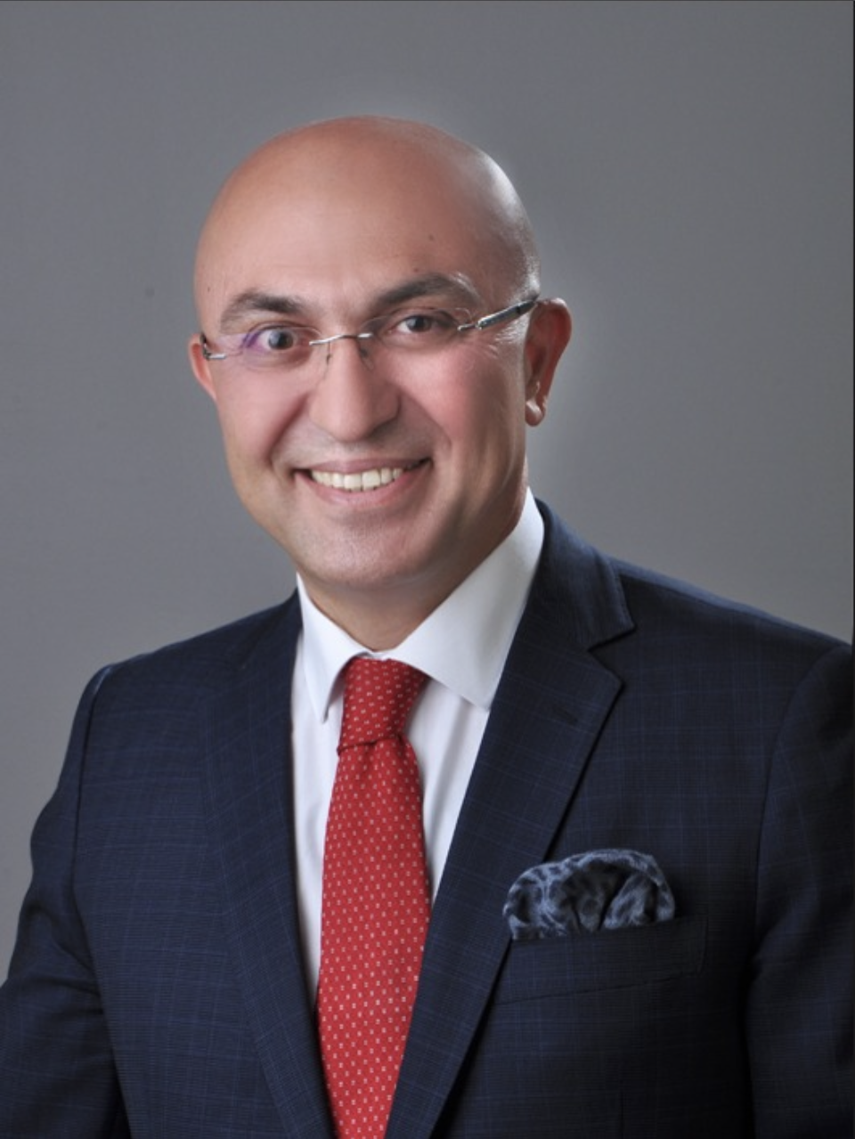 A smiling man wearing glasses, a dark checkered suit, white shirt, red tie, and a patterned pocket square.