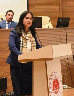 A woman speaking at a podium during a conference, with a man and a woman in the background seated at a panel.