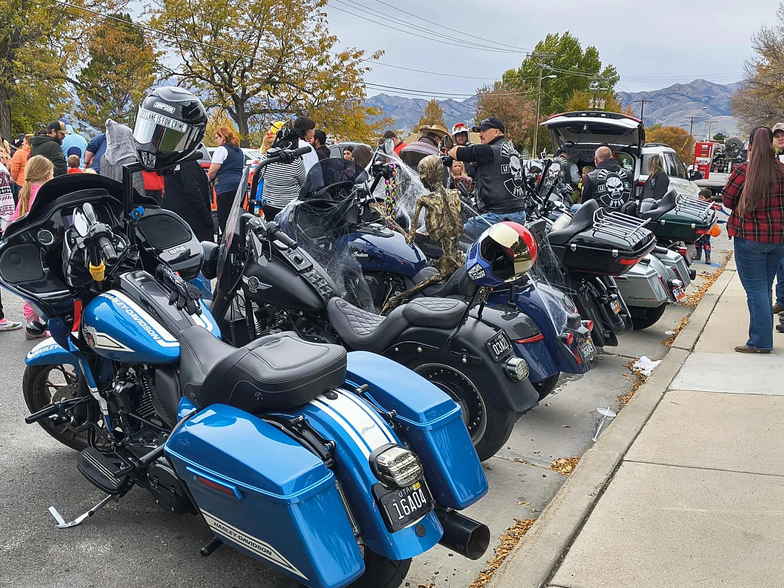 Row of parked motorcycles on a busy street during an outdoor event, with people and trees in the background and mountains in the distance.