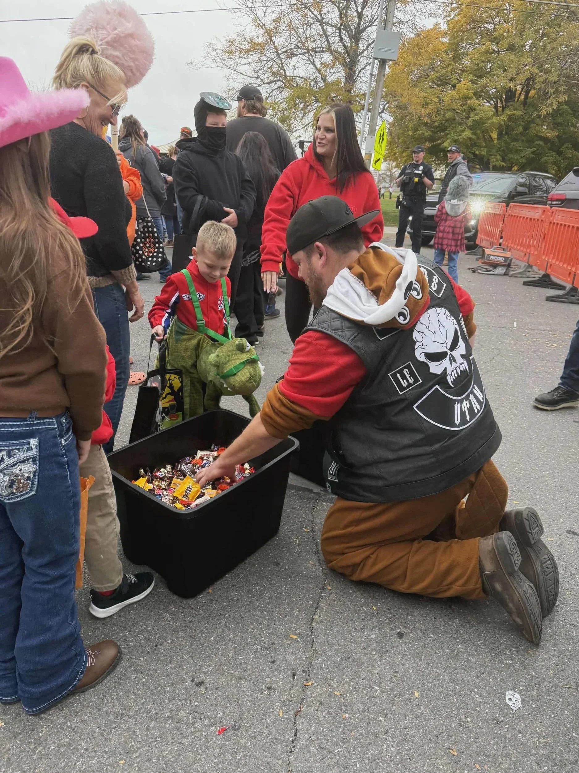 A man dressed in a leather jacket with a skull design kneels on the ground, handing out candy from a large black container to children during trick-or-treating on Halloween. The children and people surrounding are wearing costumes, with police officers and other individuals in the background.