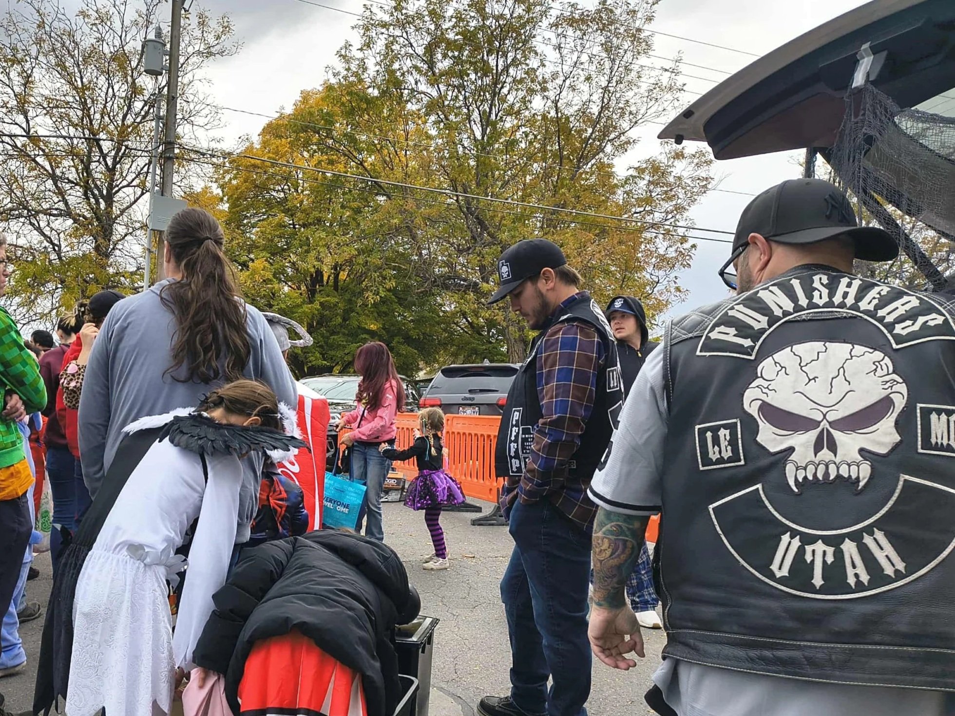 People standing outdoors on a cloudy day, some wearing biker jackets, with a child in a purple tutu, trees with fall foliage, and parked cars in the background.