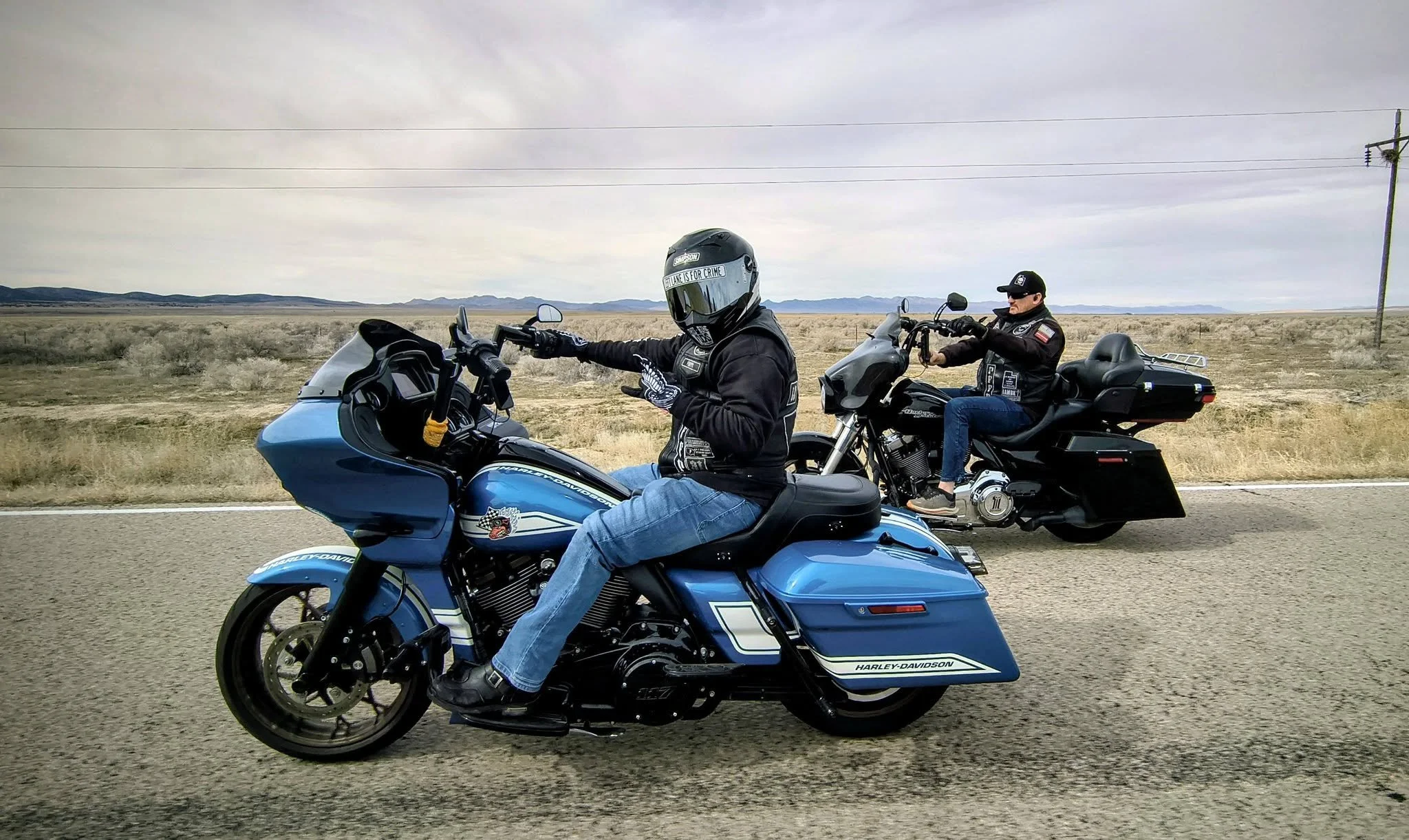 Two motorcyclists riding on a highway through a desert landscape with mountains in the background. One rider is on a blue Harley-Davidson touring bike, wearing a helmet and black jacket. The other rider is on a black Harley-Davidson touring bike, wearing a black jacket and cap.