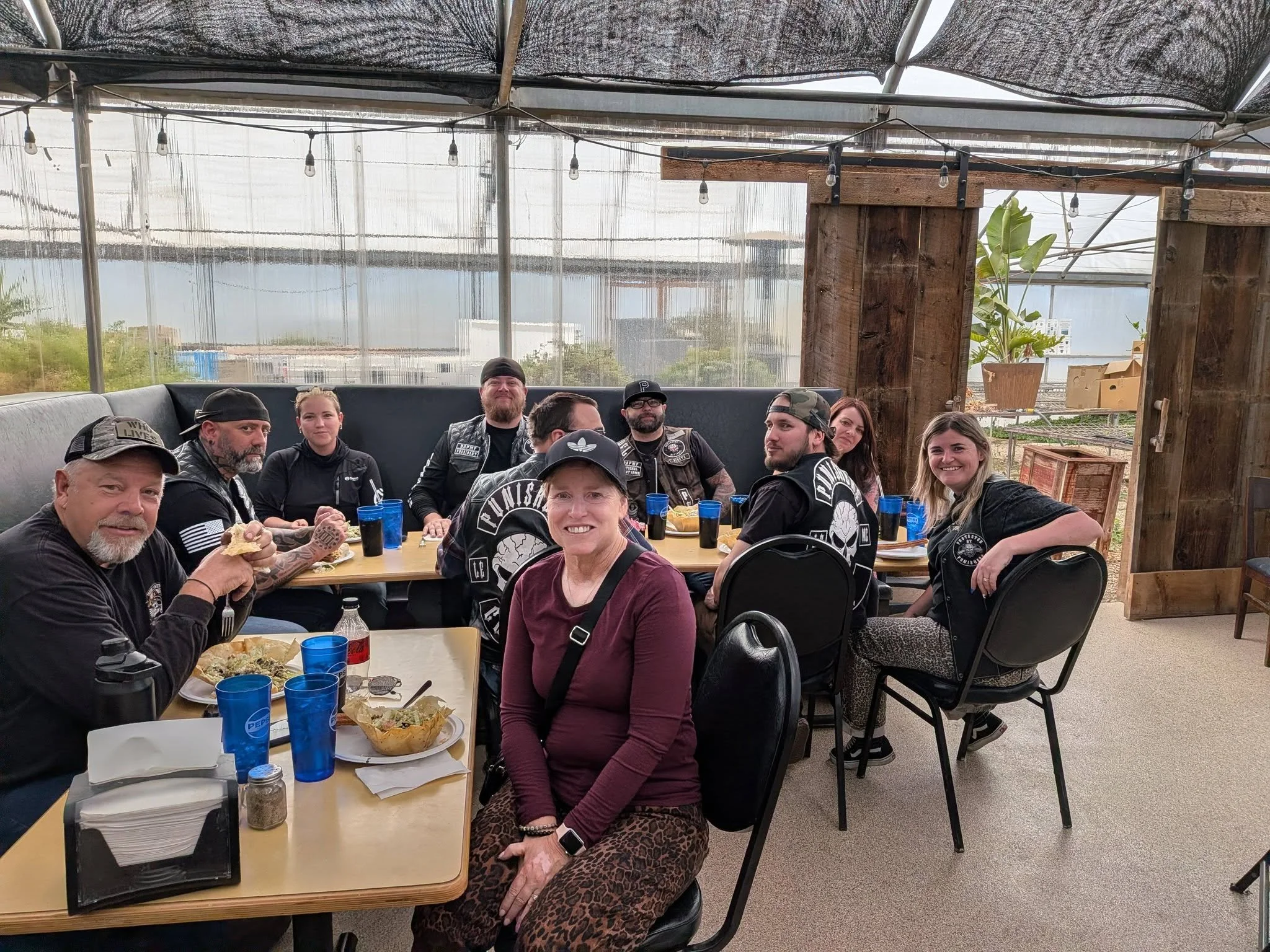 Group of people sitting at a long table in a greenhouse or covered outdoor area, enjoying a meal together.