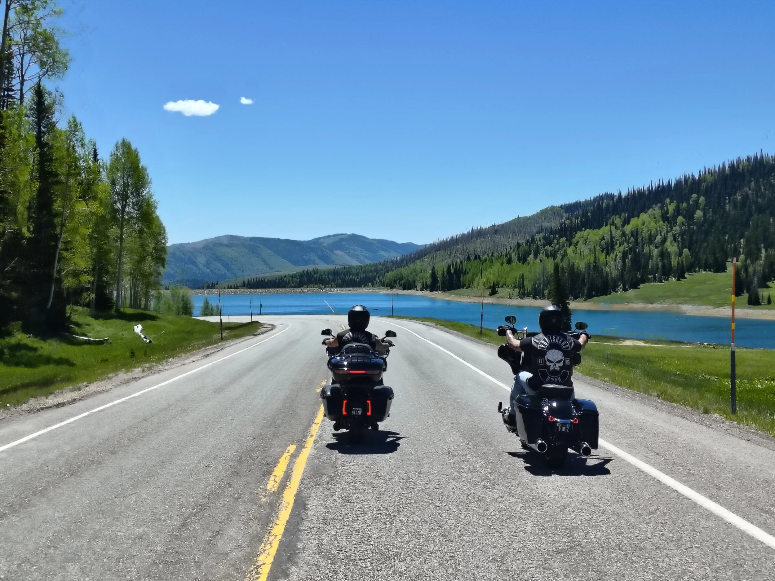 Two motorcyclists riding on a scenic mountain road next to a lake, surrounded by green trees and hills under a blue sky with a few clouds.