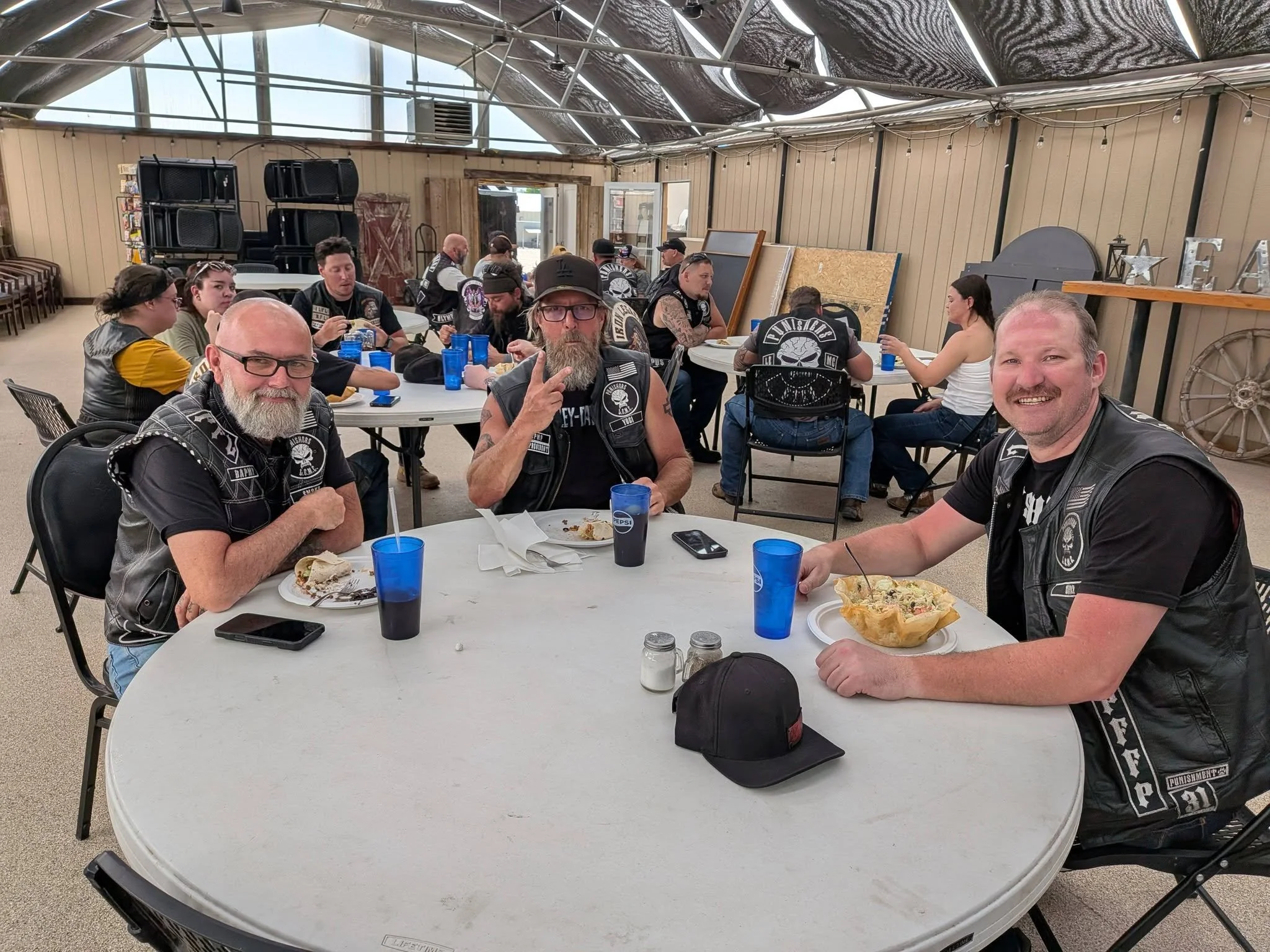 Group of people, mostly men, sitting around a table eating and socializing inside a covered patio or pavilion. Some individuals are wearing biker vests with patches and logos.