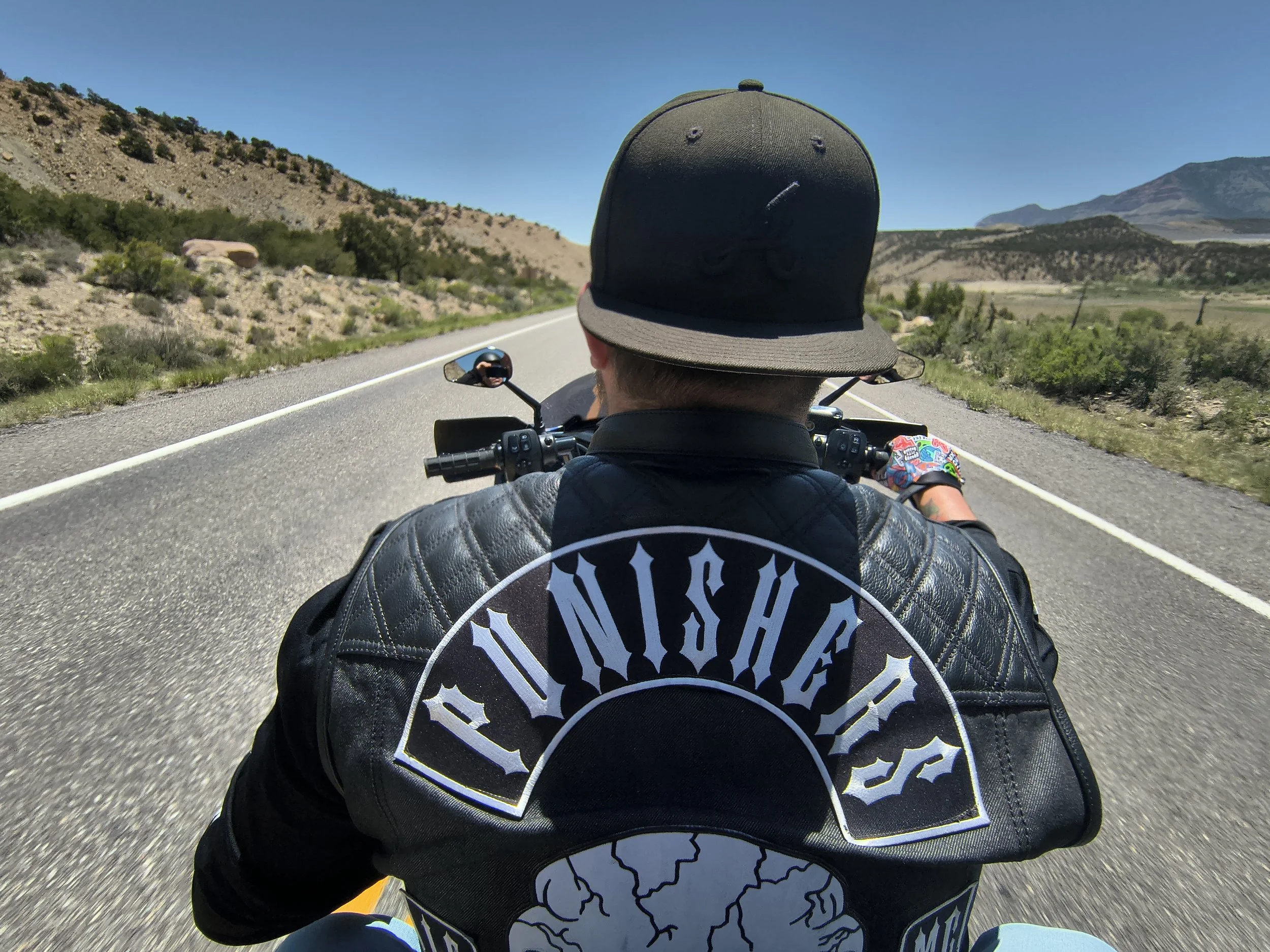 Person riding a motorcycle on a highway through a desert landscape with mountains in the background, wearing a black cap and a jacket with a graphic design.