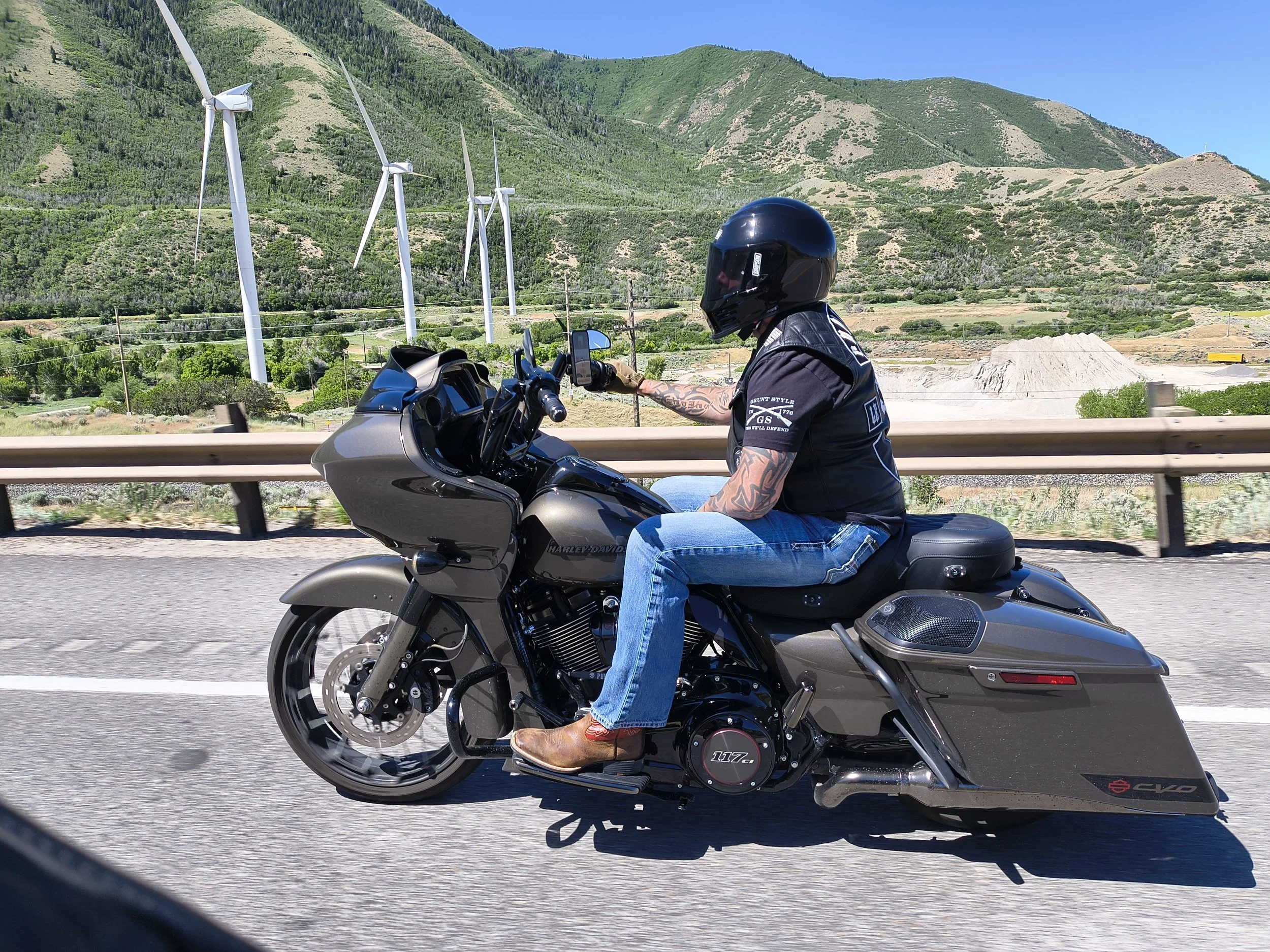 A person riding a black Harley Davidson motorcycle on a highway with mountain scenery and wind turbines in the background.