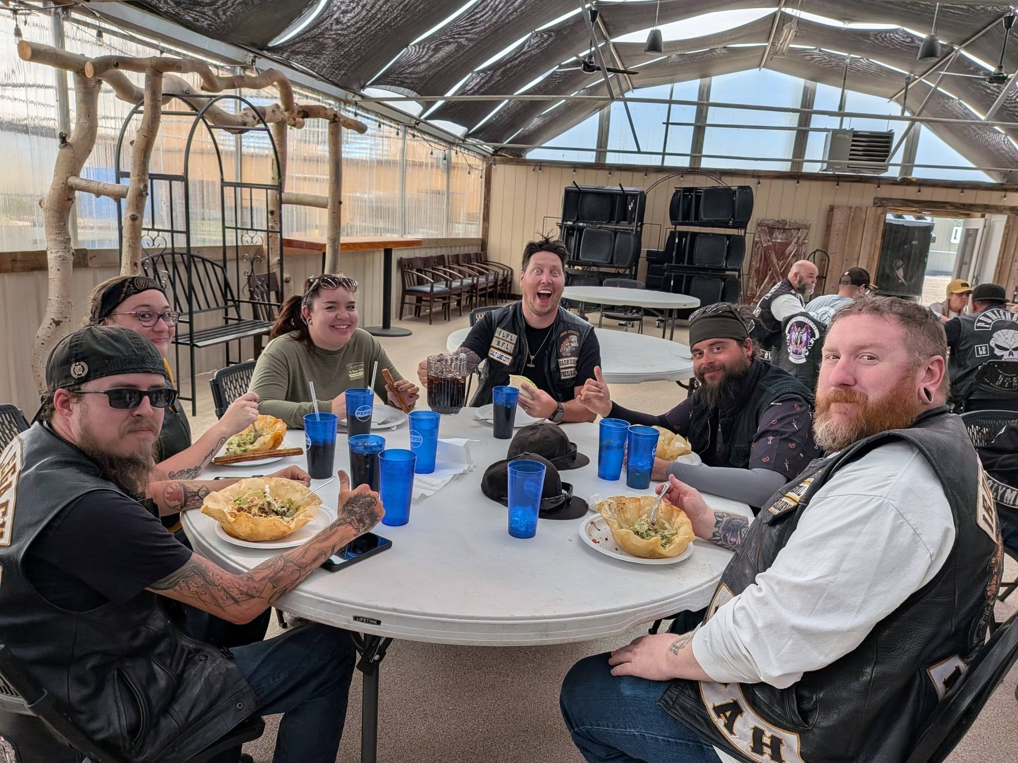 A group of people sitting around a large round table enjoying food, with some holding food and drinks, inside a rustic, partially enclosed outdoor space with a high, sloped ceiling and wooden walls, some stacked black chairs in the background.