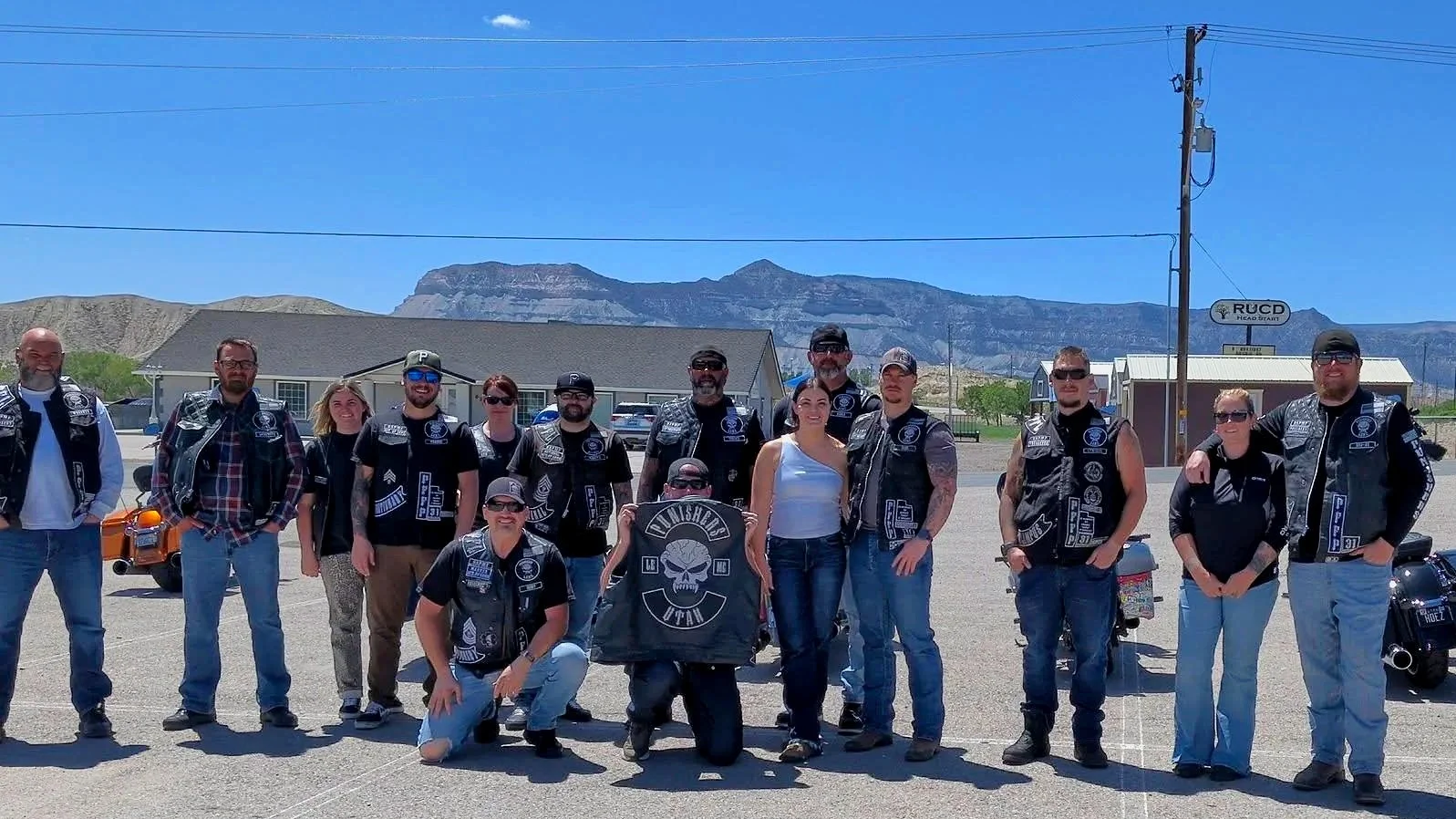 Group of motorcycle riders posing outdoors with mountains in the background and a sign that says 'RUCD'