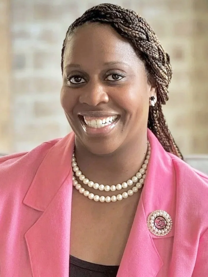 A smiling woman with dark braided hair, wearing a pink blazer, pearl necklace, pearl earrings, and a decorative brooch, standing against a blurred background.
