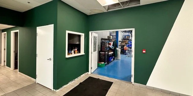Green wall with a small window and an open door leading to a storage or workshop area with shelves and blue floors.