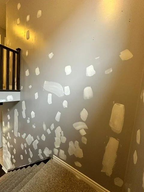 A wall with drywall patches and spackle spots, near a staircase with a wooden railing.