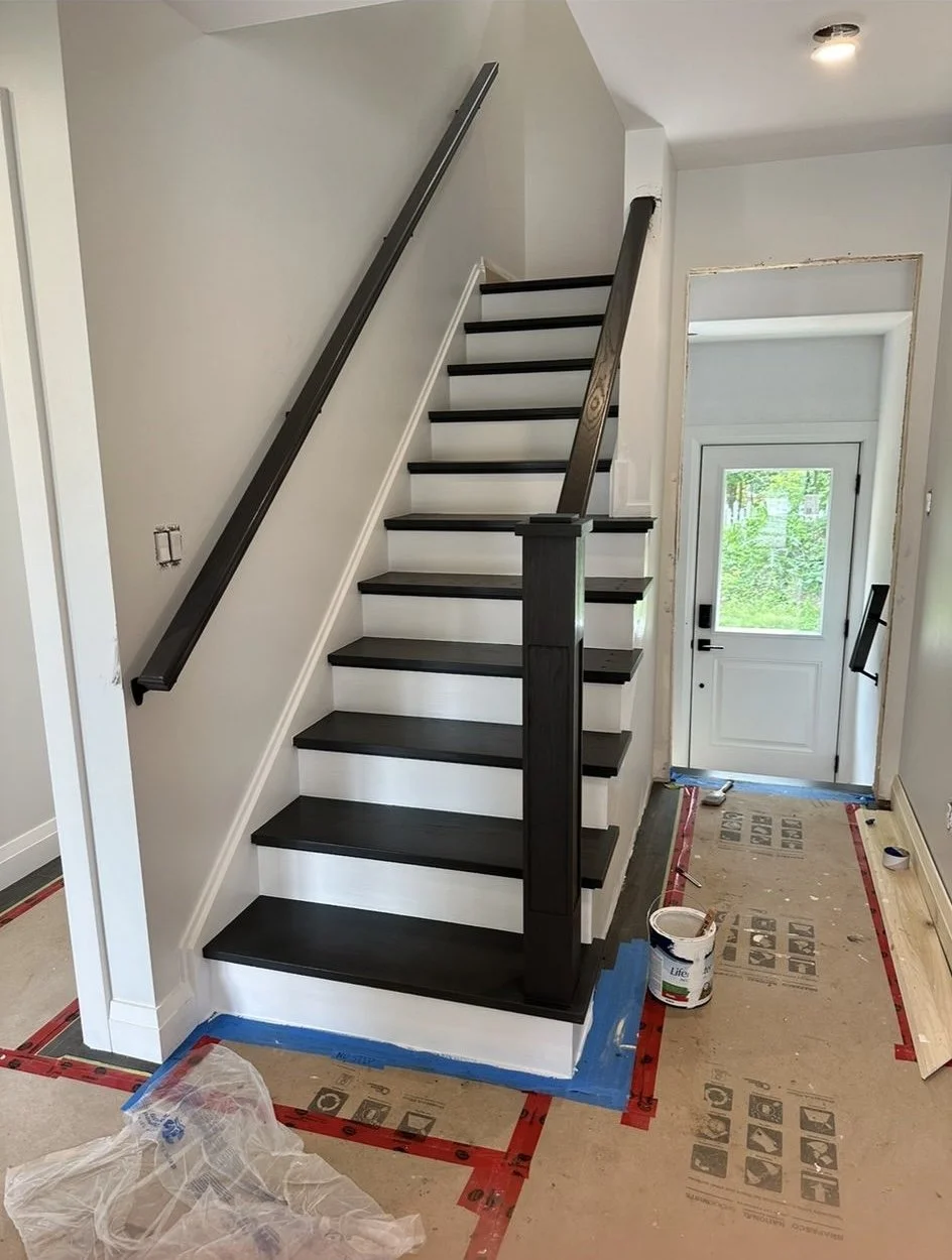 Interior of a house under renovation showing a staircase with dark wood steps, white risers, new black handrails, and unfinished walls, with construction materials on the floor.