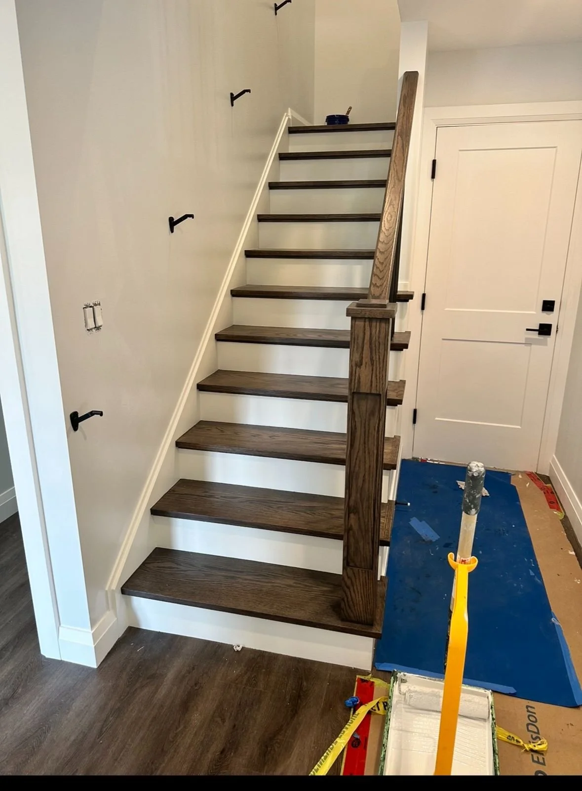 Interior view of a staircase under construction, showing dark wood steps, a partially painted white wall, and a closed white door at the top right. Construction tools and materials are on the floor, including a blue mat, a yellow level, and drywall supplies.