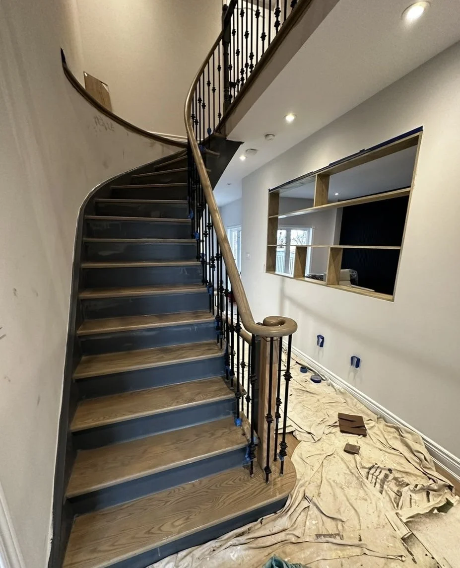 Interior of a house under renovation showing a curved staircase with wooden steps and a black metal railing, a wall with an opening for a built-in shelf, and a newspaper-covered floor with small construction materials.