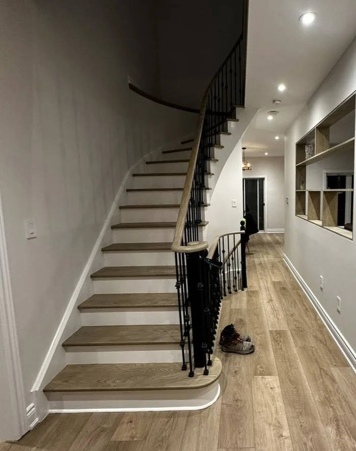 Interior view of a house with a curved staircase to the left, hardwood flooring, and white walls. There is a pair of shoes on the floor near the staircase. The hallway extends toward a closed door at the back, with built-in wall shelves on the right.