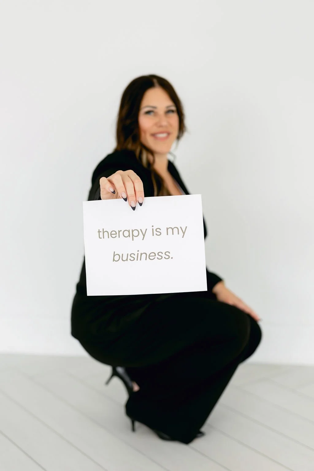 A woman crouching in black attire with a smile, holding a sign that reads "therapy is my business" against a plain white background.