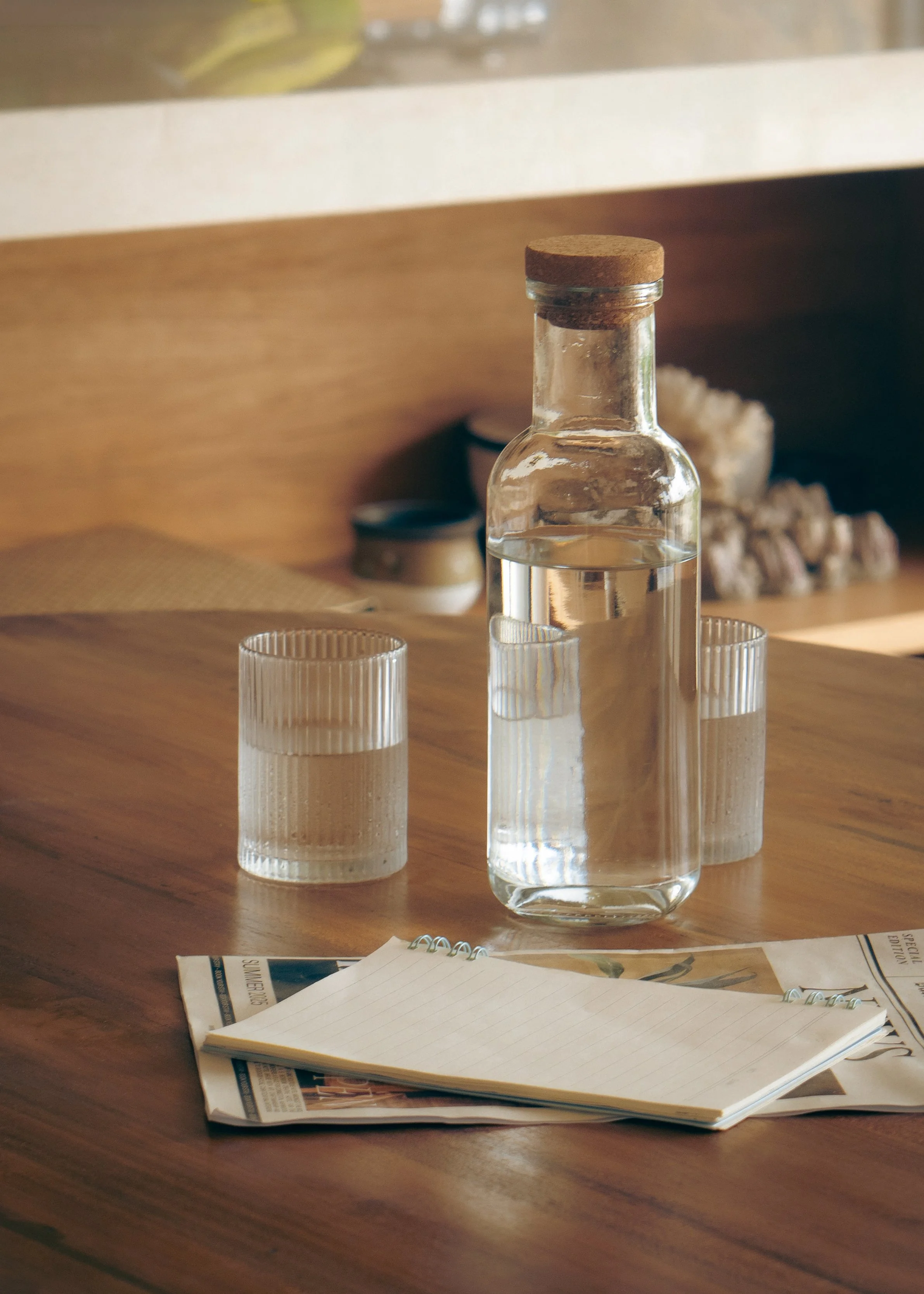 A clear glass bottle with a cork stopper, three small clear glasses, an open newspaper, and a blank lined notebook on a wooden table, with a blurred background of a shelf and decorative objects.
