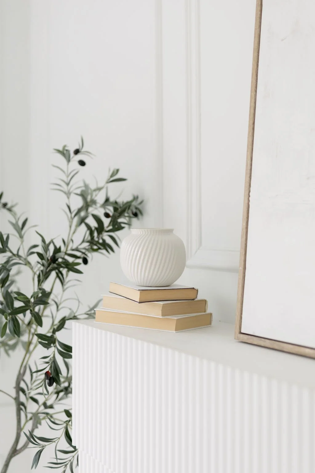 A white ceramic vase with ridges on a stack of four beige books on a white textured surface, with a plant with green leaves and black berries nearby, and a white door and blank framed artwork in the background.
