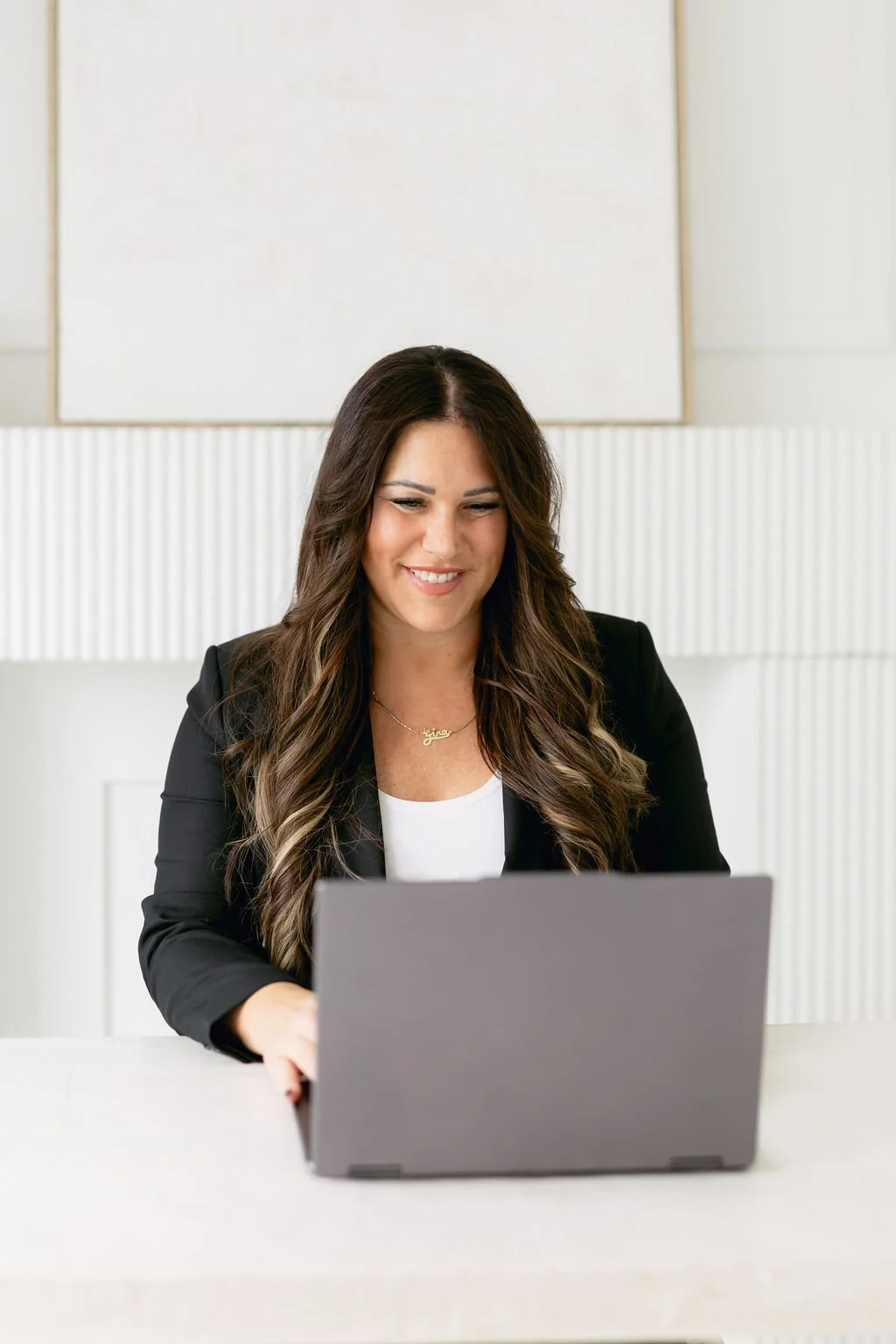 woman with long wavy brown hair working on a laptop, wearing a black blazer and white top, smiling indoors