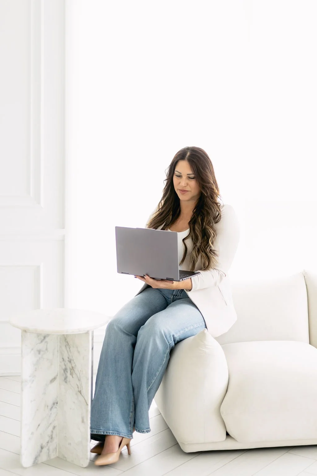 A woman sitting on a white sofa holding a laptop in her lap, in a bright white room with minimal decor.