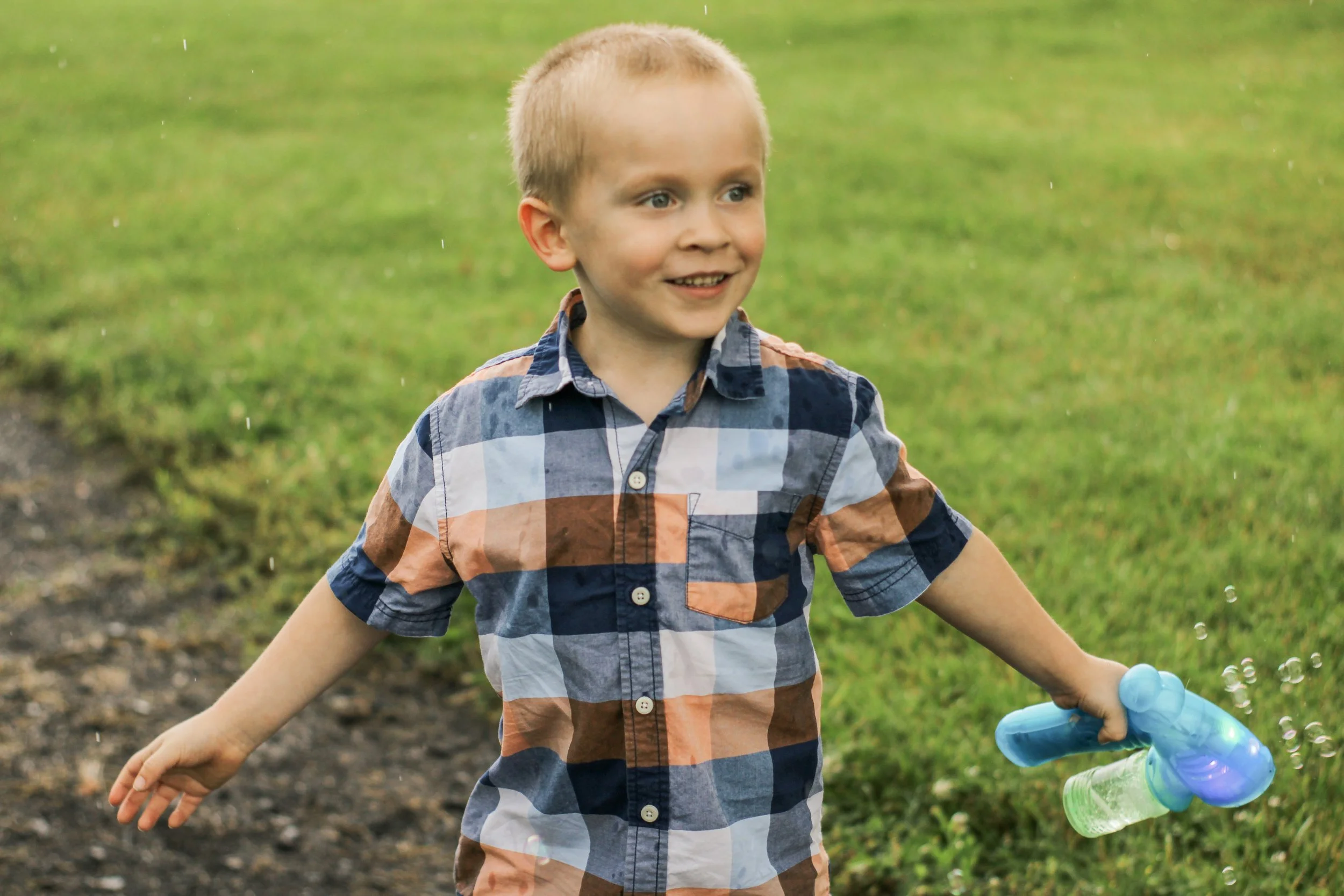 Young boy outdoors on a grassy field, holding a bubble gun, smiling, wearing a plaid shirt.