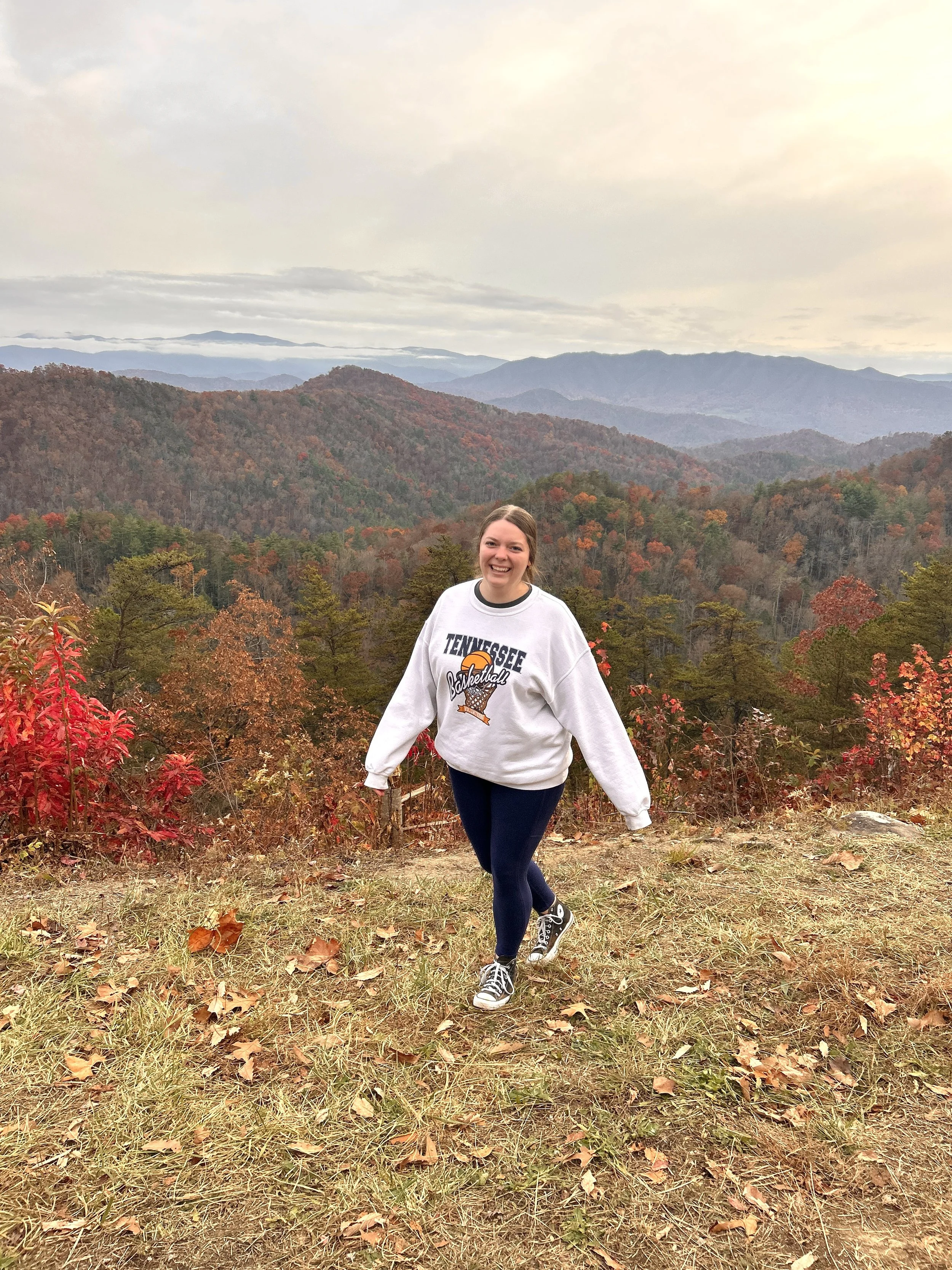 A smiling woman in a white Tennessee basketball sweatshirt, black leggings, and sneakers walking on a grassy, leaf-covered trail in a mountain landscape during autumn.