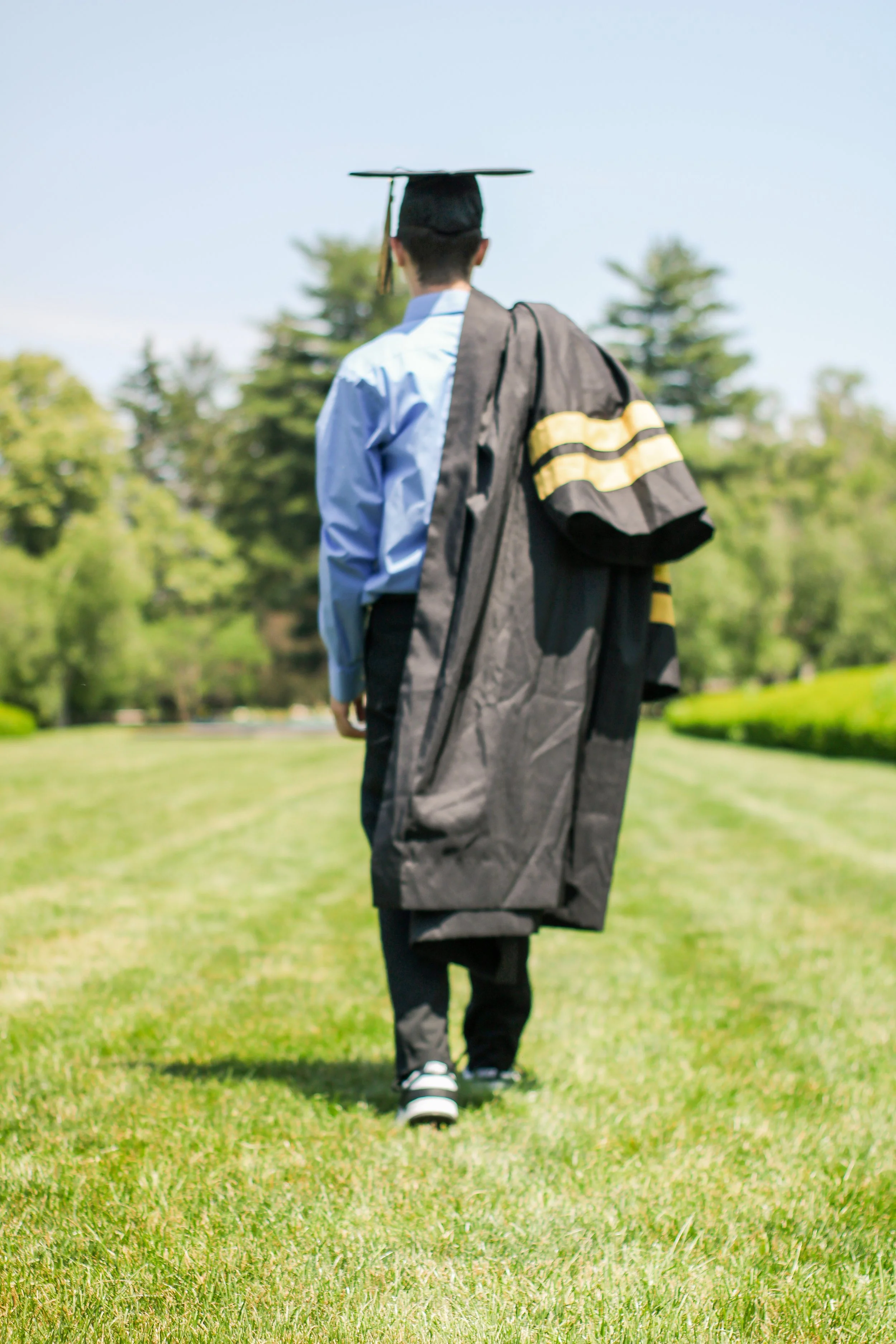 A graduate in a blue shirt and black gown, carrying a cap, walking on a grassy field with trees in the background.