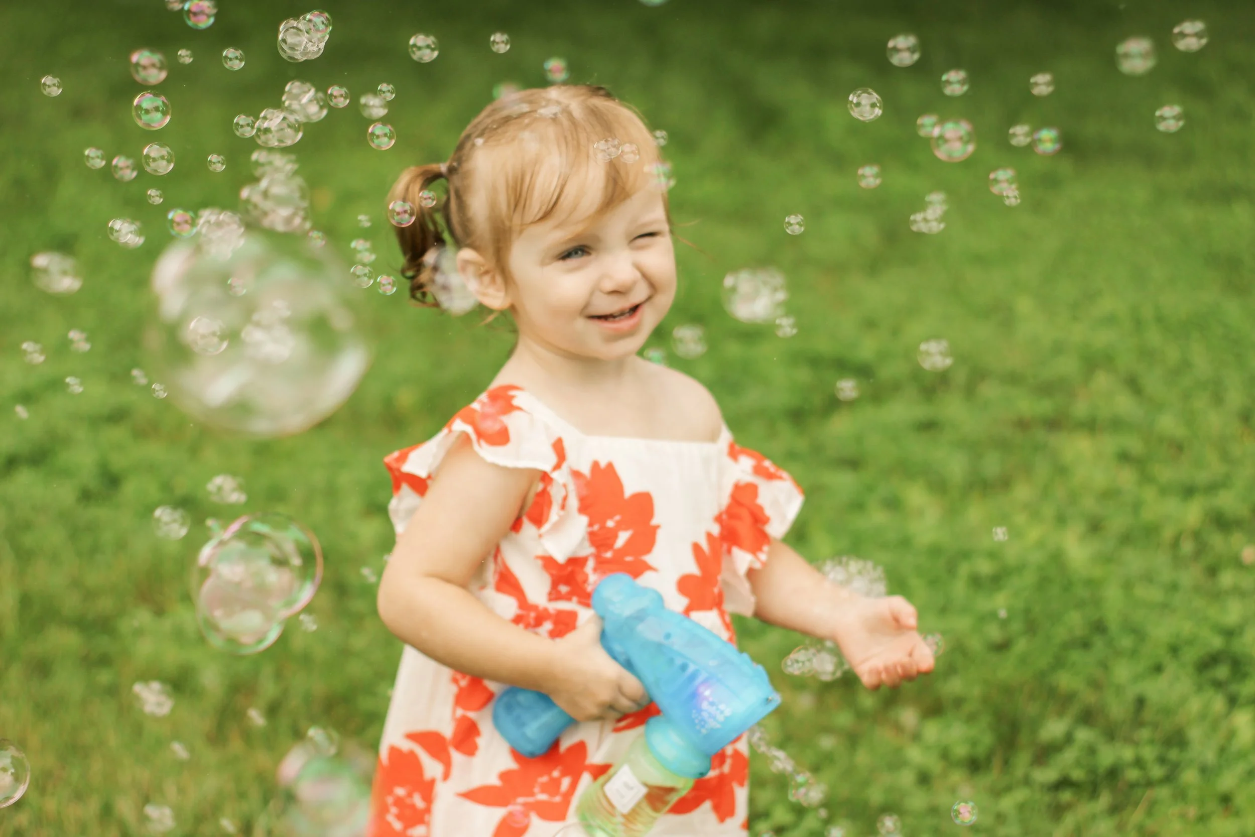 A young girl playing outdoors with bubbles, holding a bubble gun and smiling, wearing a floral dress.
