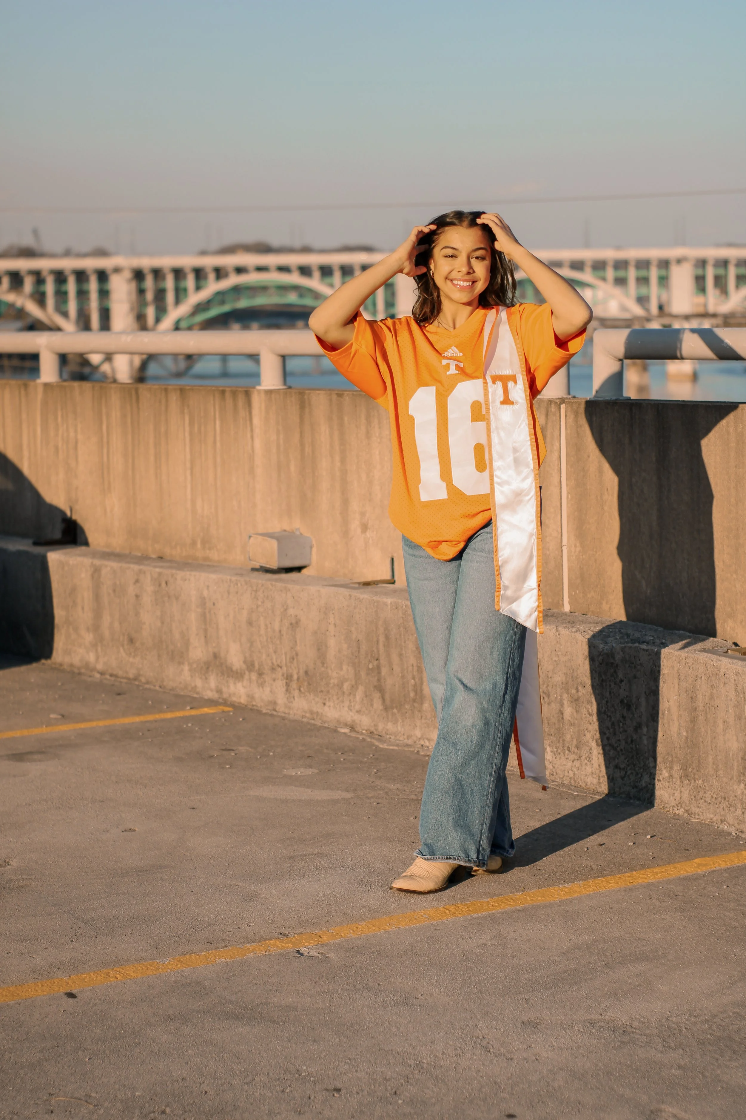 A young woman in an orange Tennessee Volunteers football jersey and jeans, smiling and posing on G10 rooftop with the Henley bridge in the background during sunset.