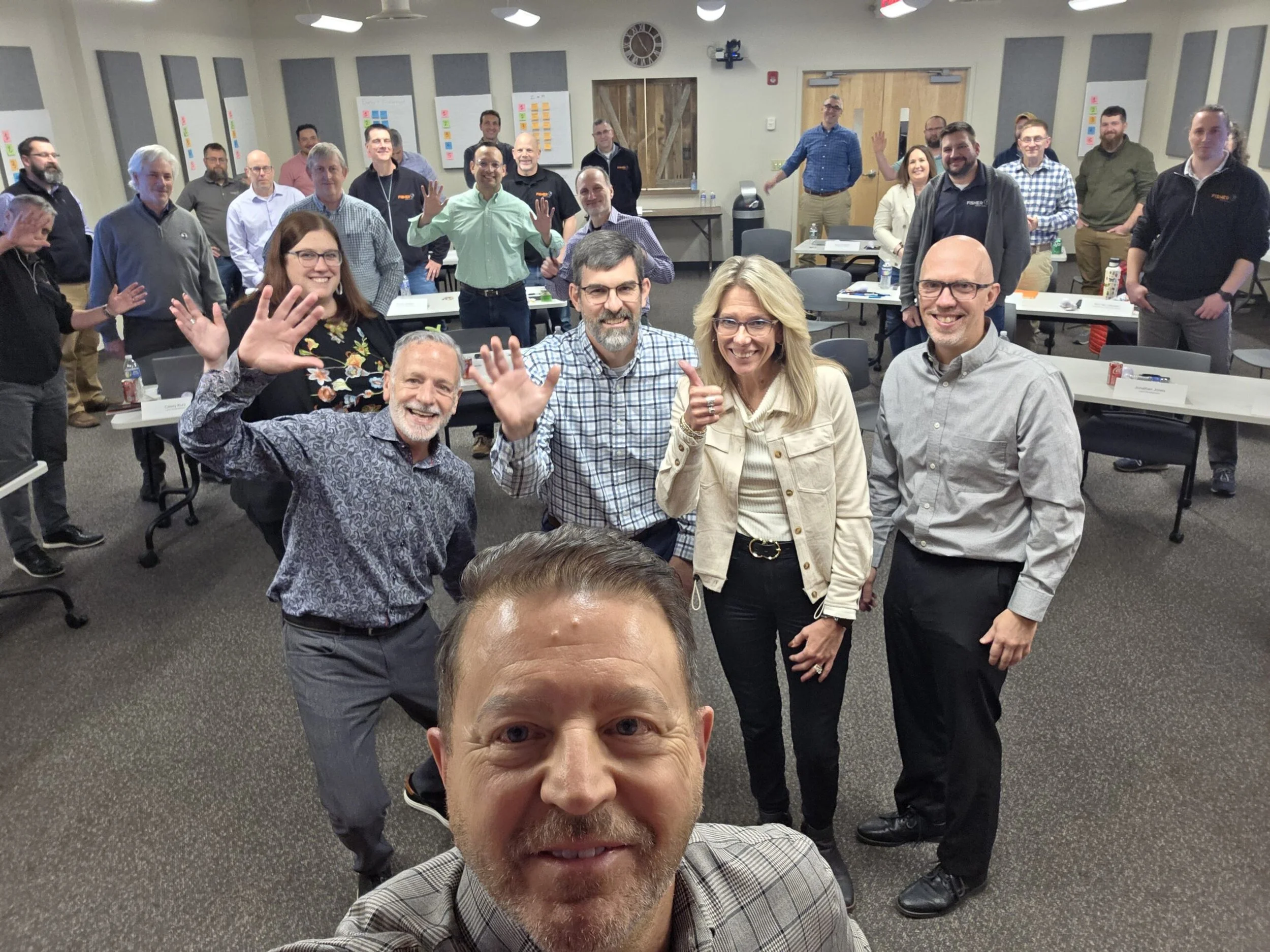 Group of people in a meeting or conference room, smiling and waving at the camera, with a presenter taking a selfie in the foreground.