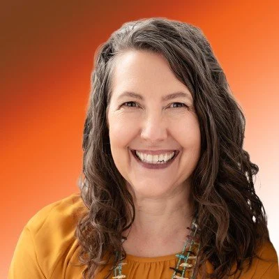 A woman with long, curly brown hair smiling in front of a red-orange gradient background, wearing a mustard yellow top and beaded necklace.
