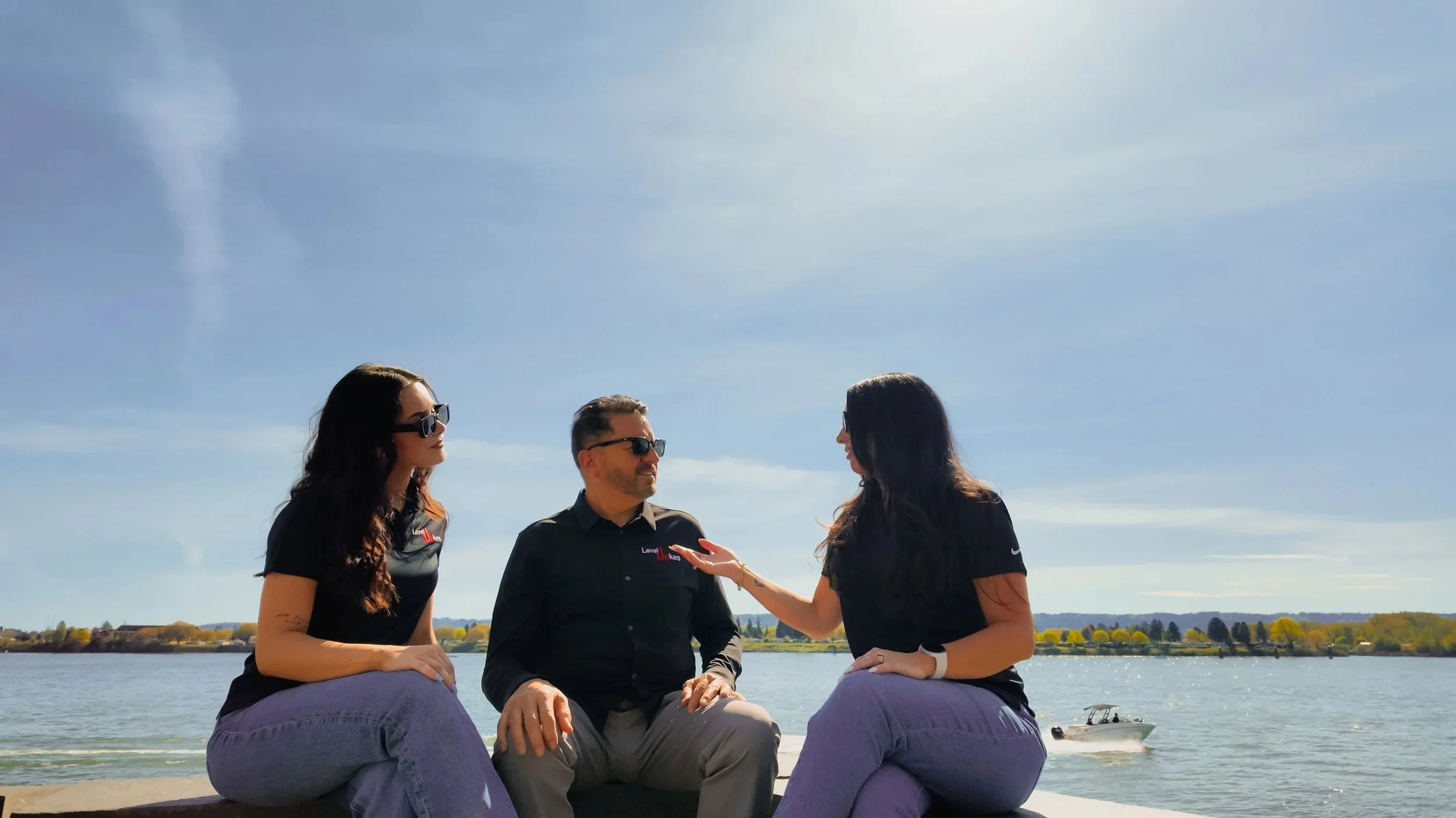 Three people sitting outdoors near a lake, engaged in conversation. Two women and one man, all wearing sunglasses, with trees and a distant shoreline in the background under a clear sky.