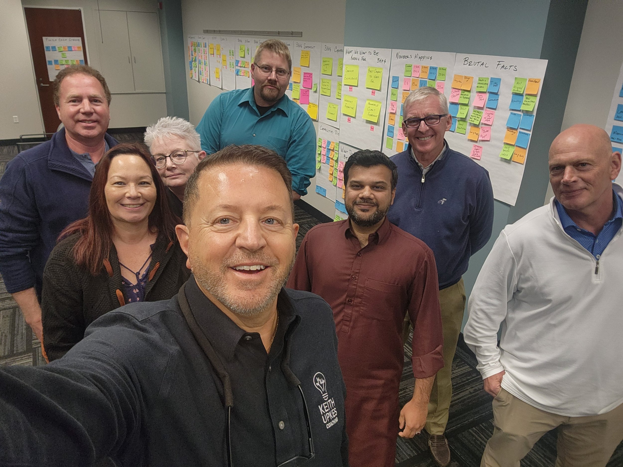 Group of eight people taking a selfie in a conference room with colorful sticky notes and charts on the whiteboards behind them.