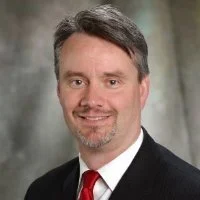 Headshot of a middle-aged man with dark hair, wearing a dark suit and a red tie, smiling against a neutral background.