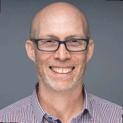 Close-up portrait of a smiling middle-aged man with glasses, short hair, and a beard, wearing a checkered shirt against a gray background.