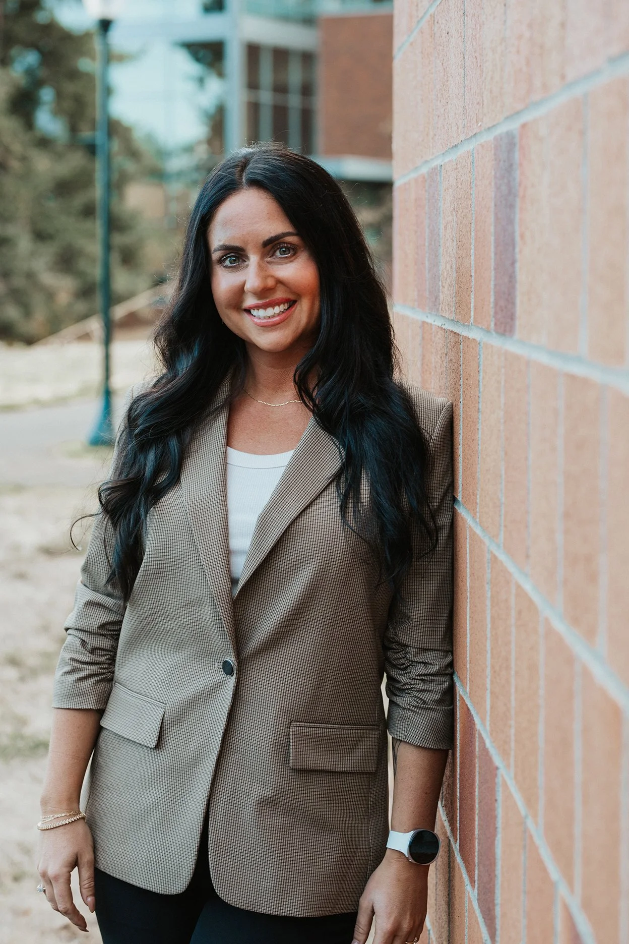 A woman with long black hair, wearing a beige blazer and black pants, smiling while leaning against a brick wall outdoors.